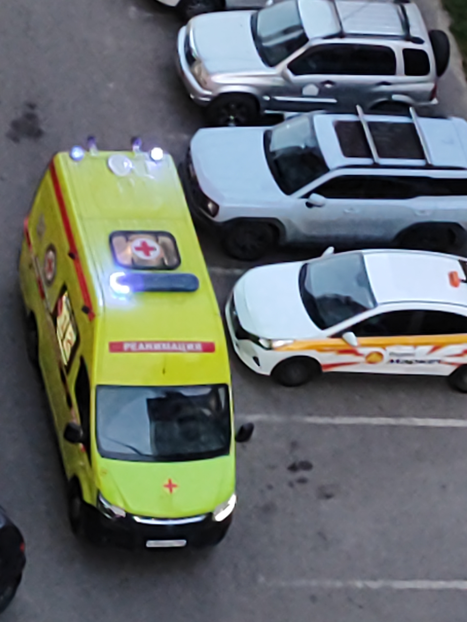 The image shows an aerial view of a parking lot with several vehicles. The most prominent vehicle is a bright yellow ambulance with red stripes and a red cross symbol on its side. It has its emergency lights flashing blue. To the right of the ambulance is a white car with a yellow and red stripe design along its side, and a logo that appears to be a stylized shell. Behind the ambulance and to its right are two SUVs, one silver and one white, parked in a lot with visible white parking lines. The ground appears to be asphalt. The overall scene suggests a parking area likely in a residential or commercial zone.