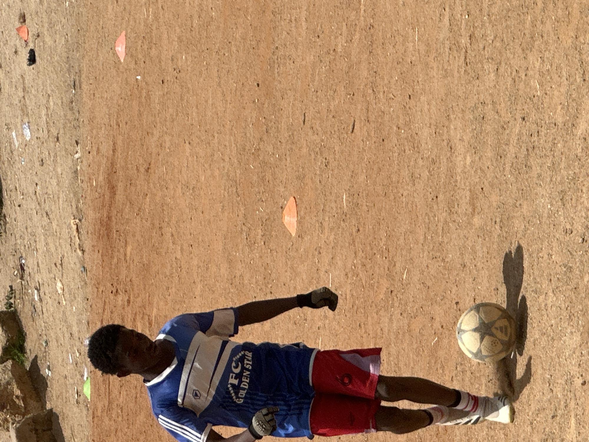 A young Black male, wearing a blue and white soccer jersey, red shorts, and white and black soccer cleats, stands on a dirt field, looking down at a soccer ball. He is wearing black goalkeeper gloves. The field is dry and dusty, with scattered rocks and debris in the background. Several orange cones are visible on the field, suggesting a training or practice session. The lighting indicates it is daytime. The scene is consistent with a casual soccer practice on an unpaved field in Jos, Nigeria.