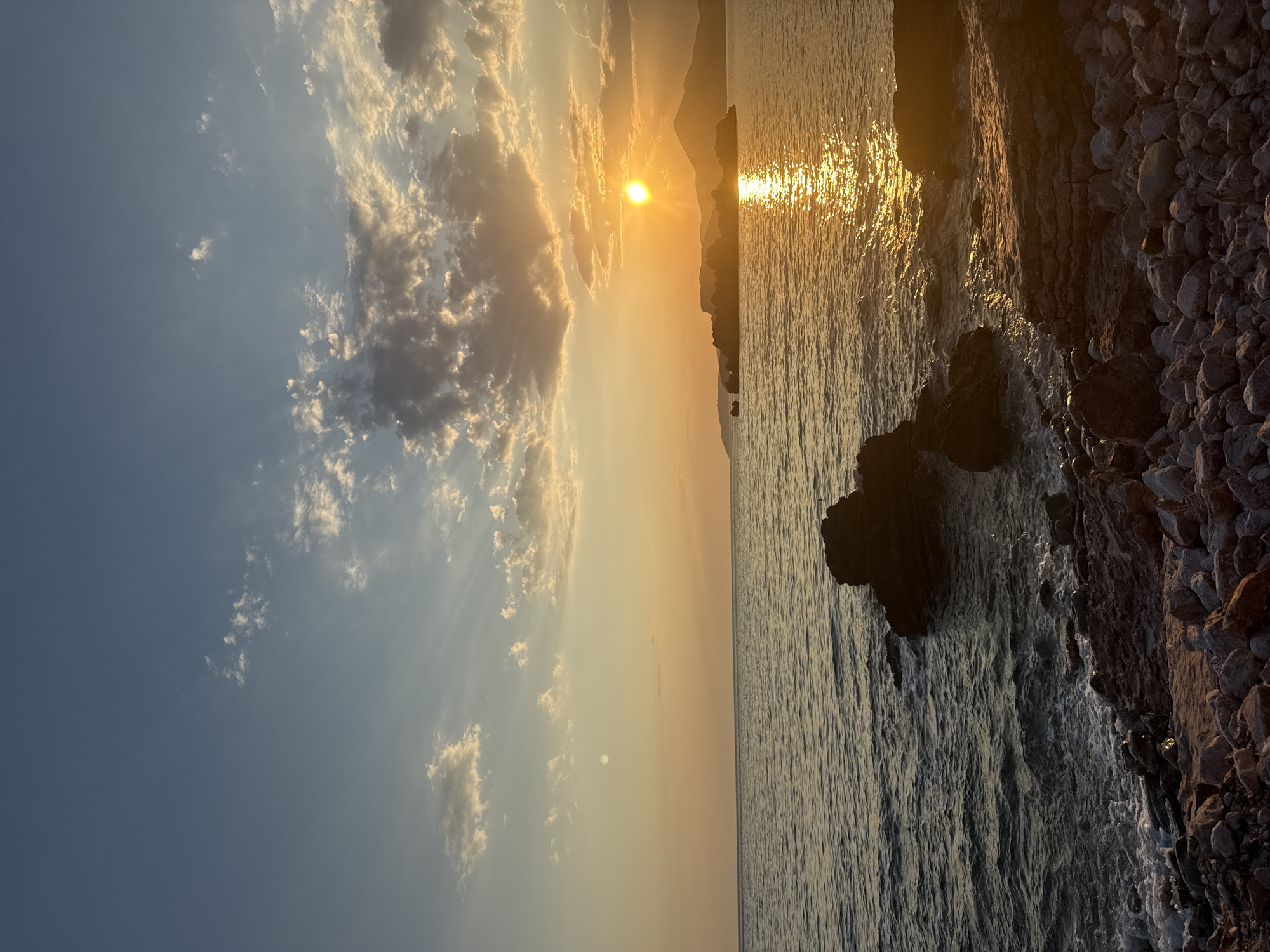 The image depicts a serene seascape at sunset in Kamenovo, Montenegro. The sun, a brilliant orb of orange and yellow, is low on the horizon, casting a golden, shimmering path across the calm waters of the Adriatic Sea. Wispy clouds, tinged with warm hues of pink and gold, drift across a pale blue sky, creating a dramatic contrast with the clear expanse above.
The foreground is dominated by a rocky shore, composed of smooth, grey pebbles and larger, dark boulders. Waves gently lap against the shore, creating white foam that contrasts with the dark, wet stones. In the distance, a silhouette of rolling hills or mountains forms the backdrop, their forms softened by the atmospheric haze and the fading light. The overall mood is one of tranquility and natural beauty, evoking a sense of peace and awe at the end of the day. The time of day is clearly sunset, indicated by the low sun and the warm, golden light. The weather appears to be clear with scattered clouds, ideal for a sunset viewing. There is no visible text in the image.