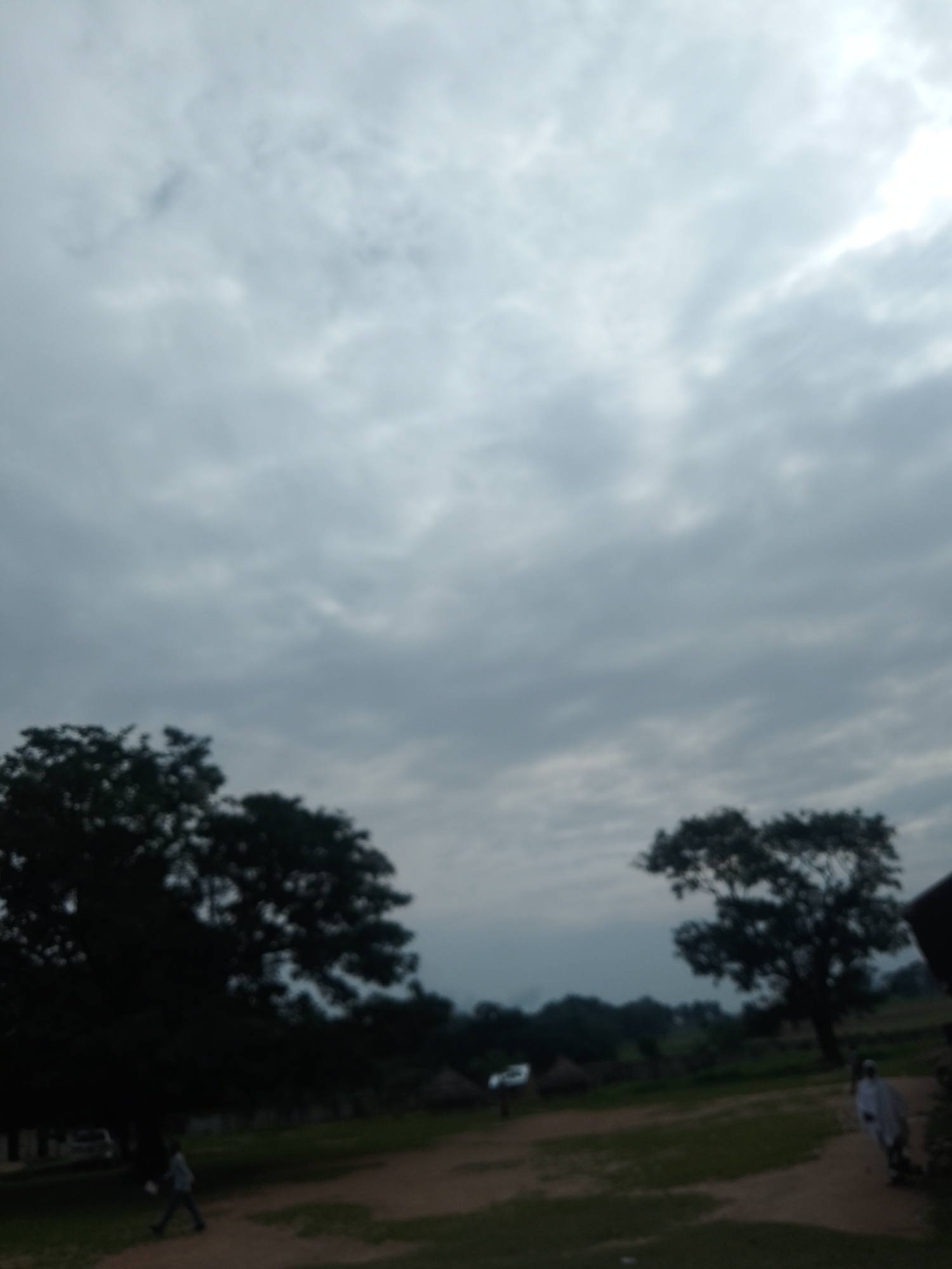 The image depicts an overcast sky with layered clouds, creating a diffused light. In the foreground, there is a dirt path with patches of green grass on either side. A few thatched-roof structures are visible in the middle ground, suggesting a rural or semi-rural setting, possibly a village in Serti, Nigeria. Two large trees with dense foliage stand out on the left and right sides of the image. In the lower left, a person is walking on the path, and in the middle right, another person in a white garment is walking away from the camera. A white vehicle is parked on the left side of the path. The overall scene is somewhat blurry and lacks sharp detail, possibly due to motion blur or low image quality.