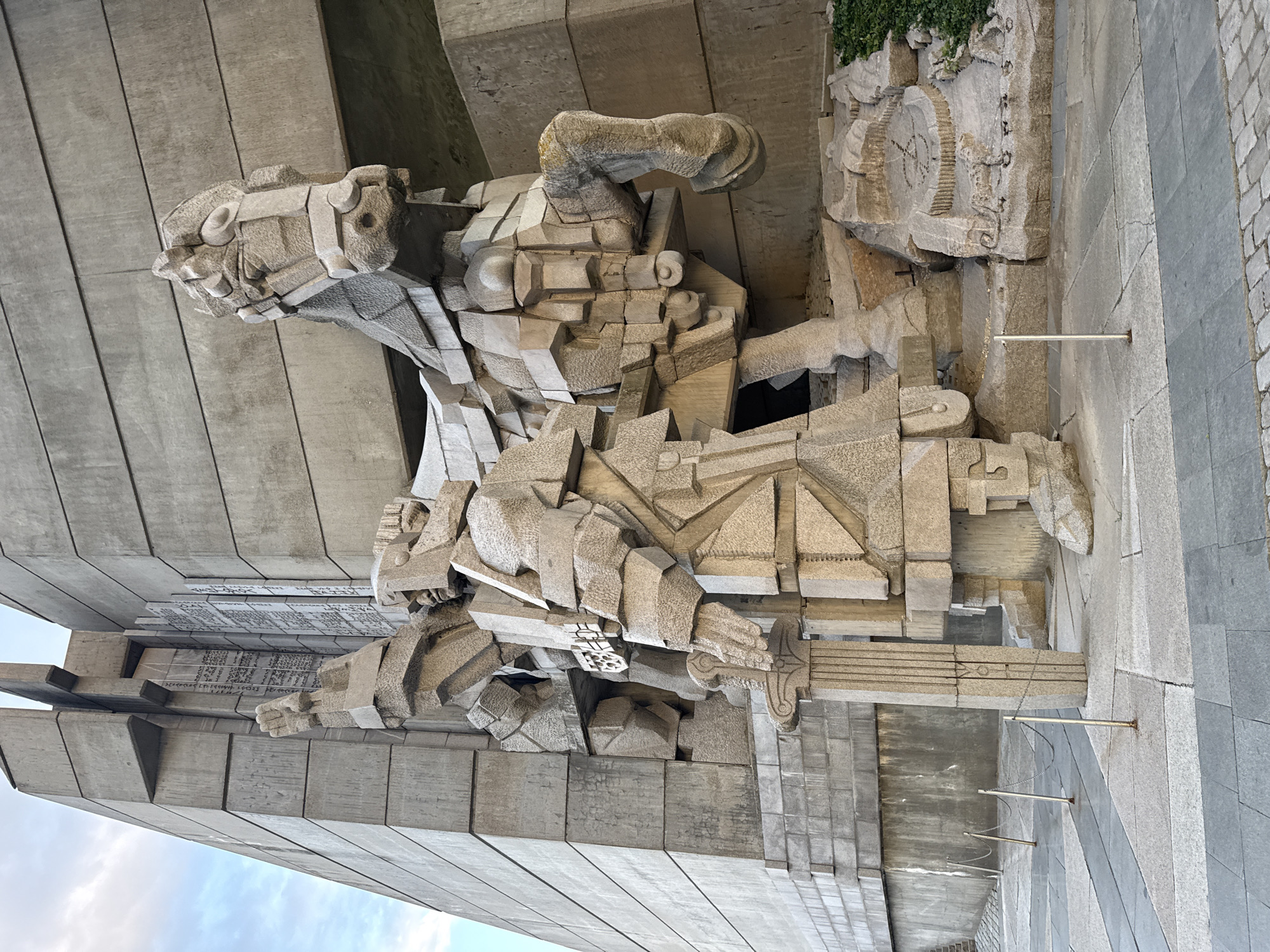 The image shows a detailed view of a socialist-era monument in Shumen, Bulgaria. The monument is constructed from large, rough-cut stone blocks, creating angular depictions of figures in a dynamic, heroic style. Two figures dominate the scene, appearing to be warriors or historical figures, their forms abstracted and fragmented. In the background, there are flat concrete slabs inscribed with text, possibly names or historical information. To the right, a stone structure depicts a clock-like design. Metal poles and chains are visible in the foreground, possibly serving as barriers. The setting appears to be an outdoor public space, likely part of the monument complex.