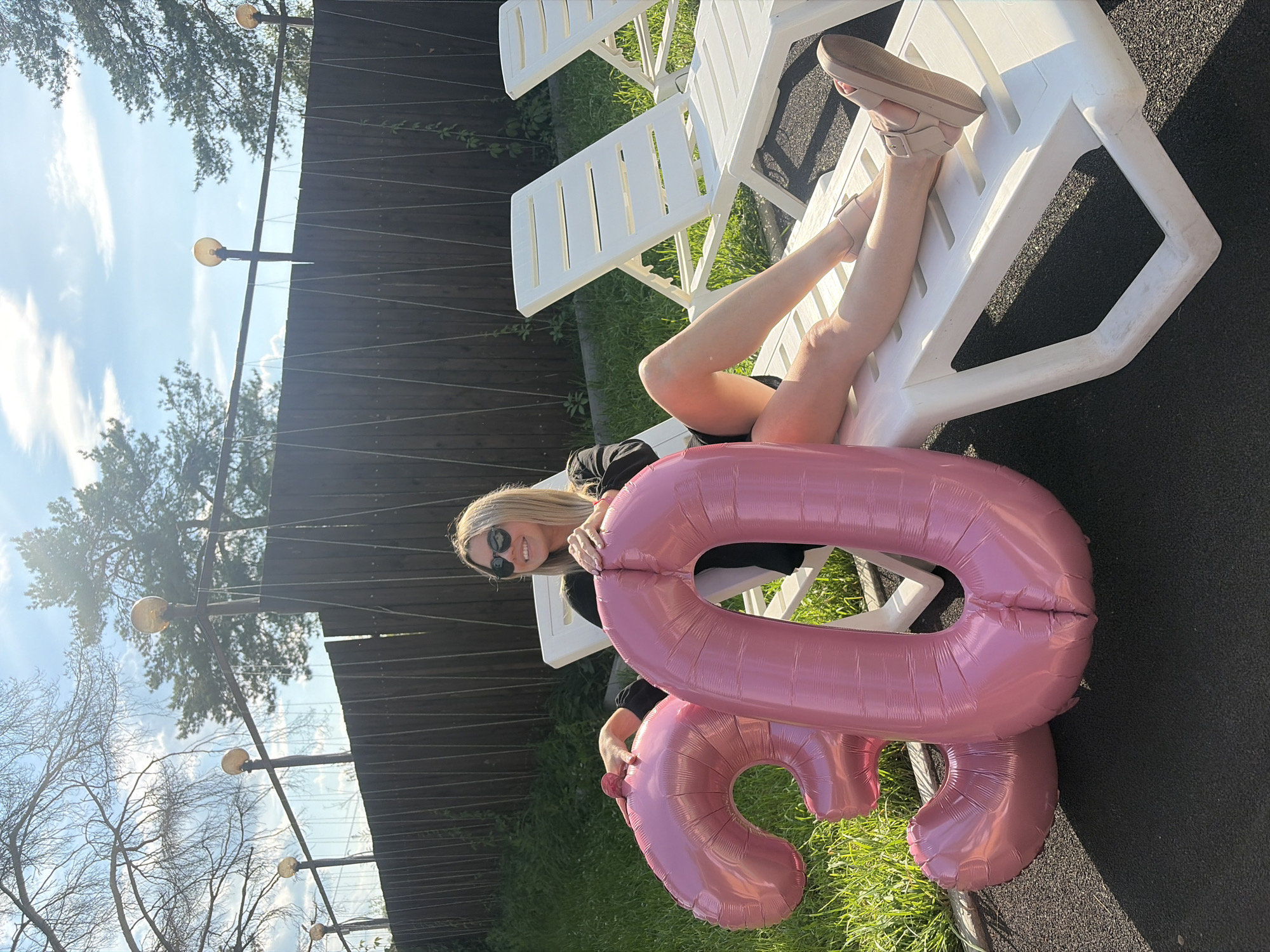 In Chaikovsky, Russia, a blonde woman is pictured outdoors, reclining on a white plastic lounge chair. She is wearing sunglasses and a black top, with her legs crossed at the ankles. Two large, pink, inflatable balloons, shaped like the numbers "3" and "0," are positioned in front of her. The balloons are partially obscuring her lower body. The background consists of a wooden fence, green grass, and trees under a bright, partly cloudy sky. The scene appears to be a backyard or a recreational area, possibly for a birthday celebration.