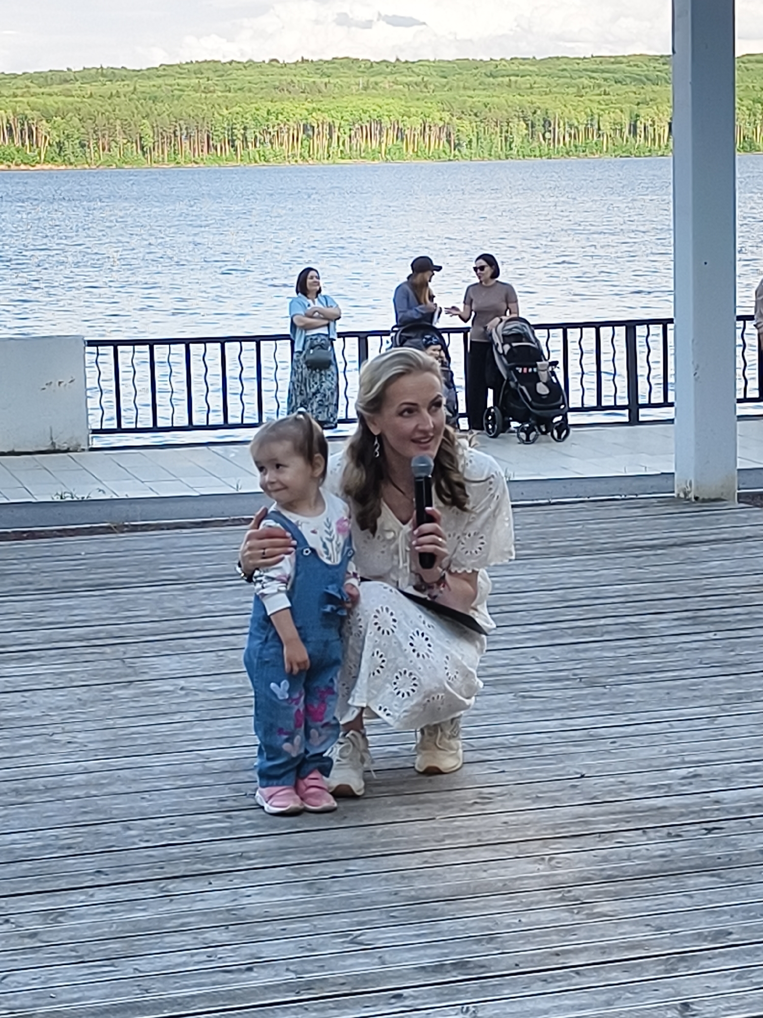 In the city of Chaikovsky, Russia, a woman kneels on a wooden boardwalk, holding a microphone in her right hand and her left arm around a young child. The woman, with blonde hair, wears a white, intricately patterned dress. The child, wearing blue denim overalls and a white shirt with a floral print, stands beside her. The child has a ponytail and looks off to the left. In the background, a large body of water, likely a river or lake, is visible, with a tree-covered shoreline. Several other people, including two women and a person pushing a stroller, stand further back on a raised platform behind a black railing. The overall setting appears to be a scenic outdoor venue near the water.