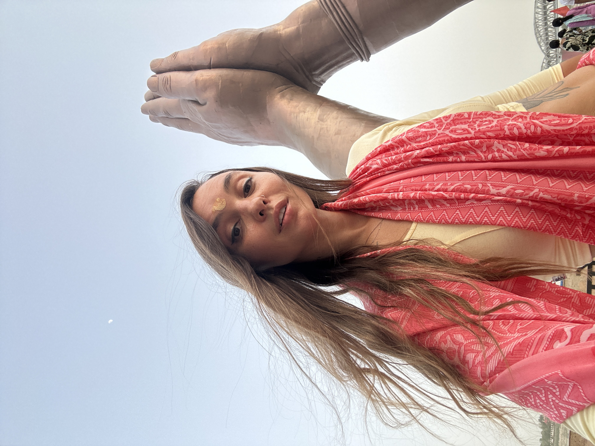 A low-angle, medium shot depicts a young woman with long, light brown hair wearing a pale yellow top and a bright pink patterned scarf. She has a gold-colored mark on her forehead. She is positioned below a large statue of clasped hands made of a material that appears to be bronze or a similar metal. The background features a pale blue sky with a small, distant object that might be a cloud or the moon. To the upper right, indistinct objects that could be structures or foliage are visible. The statue appears to be part of the Isha Foundation's Sadhguru Sannidhi statue of Adiyogi Shiva in Mughalsarai, India.
