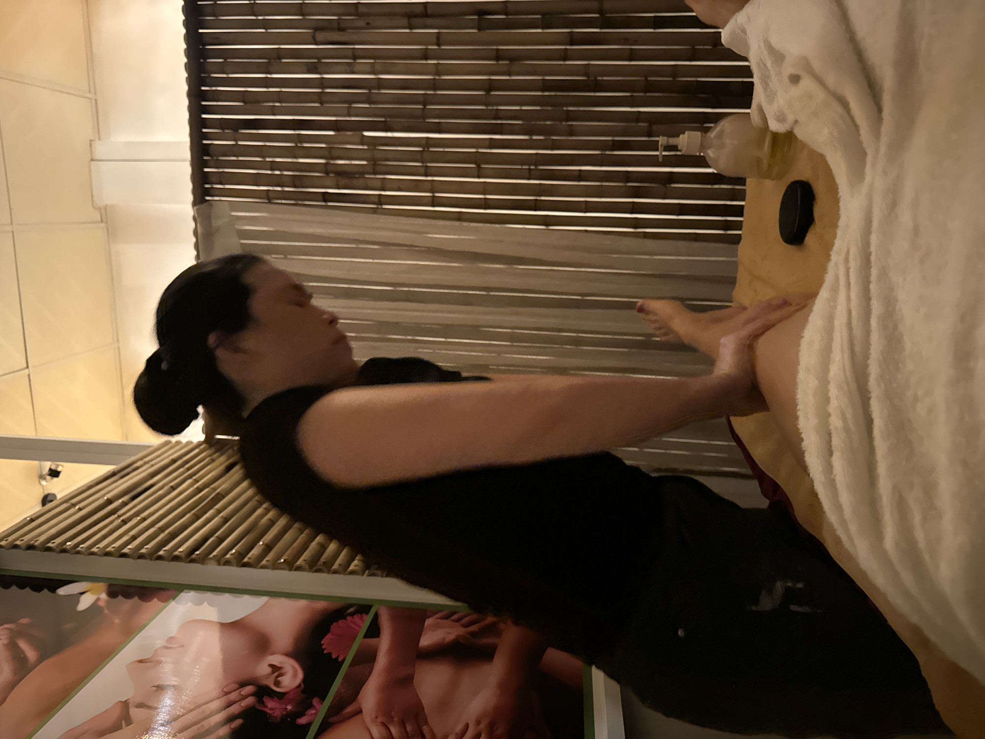 A woman receives a massage in a dimly lit room, possibly a spa, with bamboo paneling as a backdrop. She is lying on a massage table, covered by white towels. A massage therapist, wearing a black shirt and pants, is seen applying pressure to her leg. A bottle of massage oil and a black stone are visible on the table. The overall atmosphere suggests a relaxing spa experience. The image is a photograph taken indoors, and the lighting conditions are soft, creating a warm ambiance.