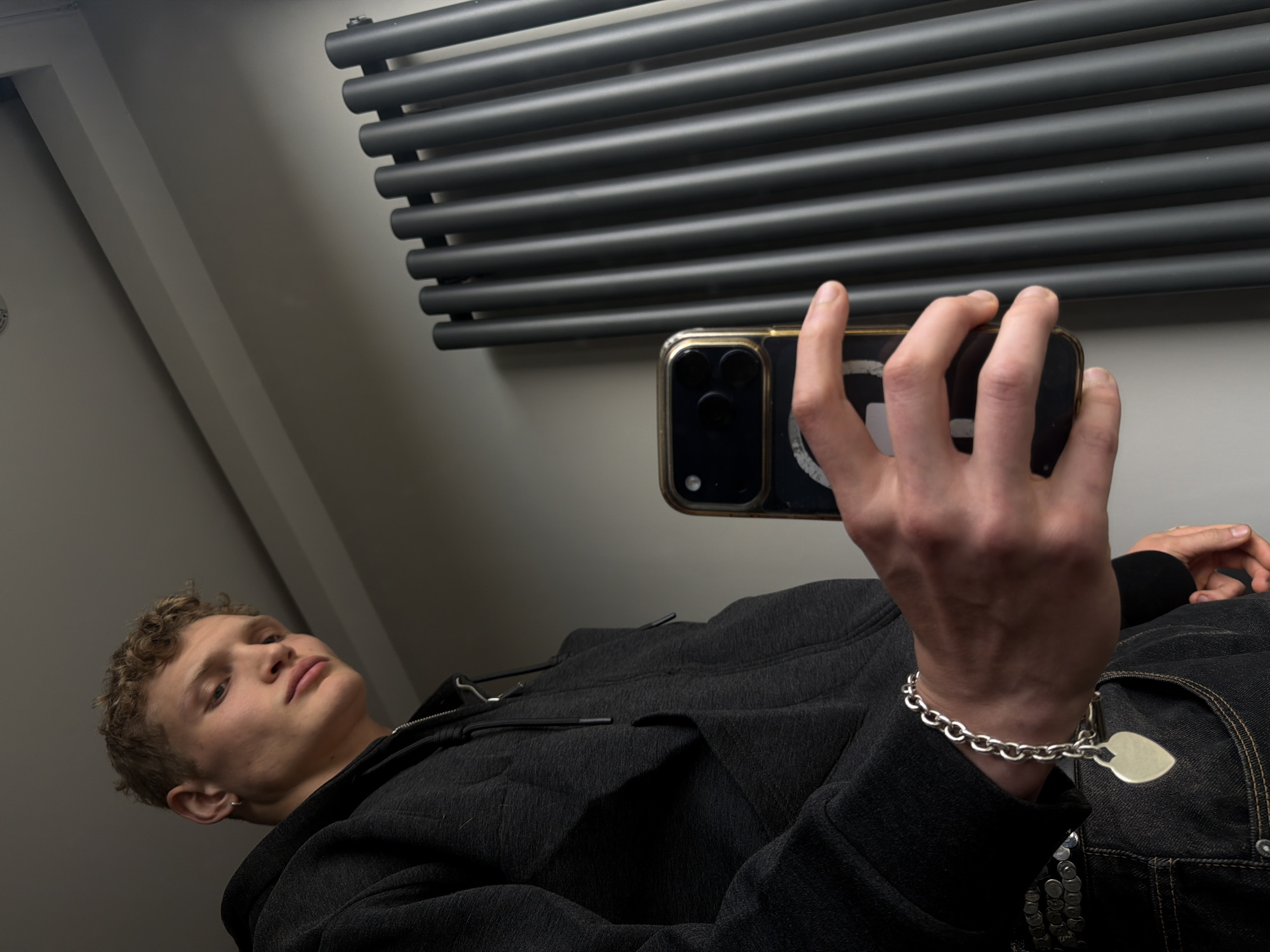 A young Caucasian male lies at an angle, appearing to be in a residential setting. He is holding a smartphone with a gold-colored case, presumably taking a photo or video. The phone's screen displays a circular white graphic. The man is wearing a dark hooded sweatshirt and jeans. A silver bracelet adorns his wrist. A gray radiator is mounted on the wall behind him. The location is indeterminable.