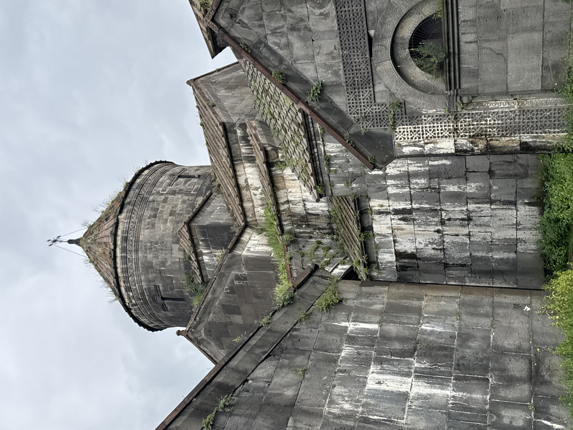The image shows a close-up, low-angle view of an ancient stone building, likely a church or monastery, with a cloudy sky as the background. The architecture is characterized by large, weathered stone blocks, a conical dome topped with a cross, and several pitched roofs covered in aged tiles. Greenery, including moss and small plants, grows in the crevices and on the roofs, indicating age and exposure to the elements. Intricate stone carvings are visible on one of the facades, featuring geometric patterns and a semi-circular arched window. The overall impression is one of historical significance and a sense of enduring presence within its environment. The location is identified as Haghpat, Armenia. No people or animals are present in the image, and no actions or interactions are depicted. The scene is static and focuses solely on the architectural elements of the building and its immediate surroundings, which appear to be overgrown with vegetation.