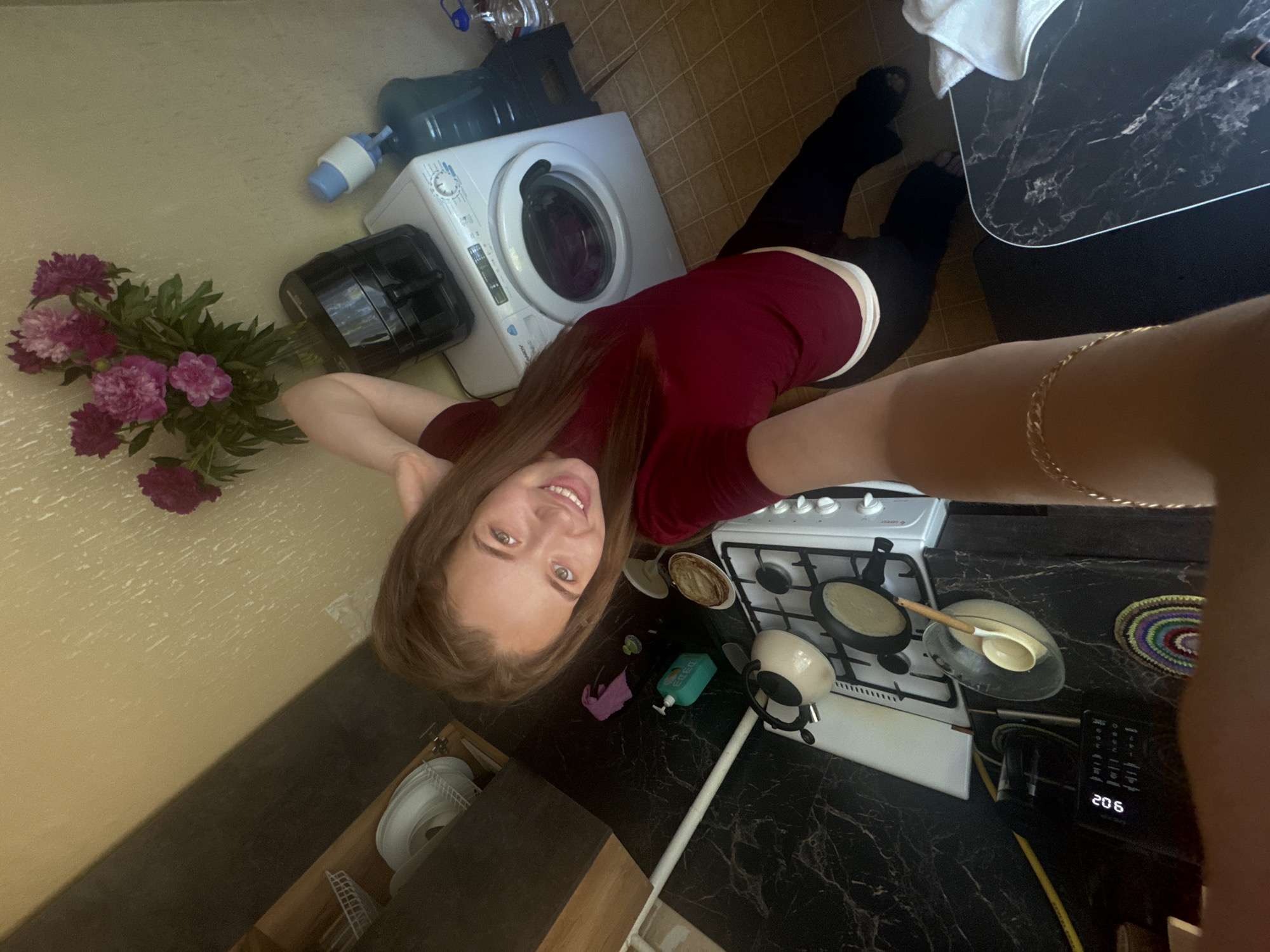 The image depicts an overhead, wide-angle selfie of a young woman in a kitchen, possibly in the city of Chaikovsky, Russia. The woman, with long brown hair and a bright smile, is wearing a burgundy t-shirt and black pants. She is positioned in the center-left of the frame, with her arms raised and her right hand holding the camera.
The kitchen environment includes a black marble countertop with a white gas stove. On the stove, there are two pans, one of which appears to be cooking pancakes, and a kettle. A glass bowl with a ladle sits on the counter next to the stove. Adjacent to the counter is a washing machine, and on top of it, a black air fryer. Behind these appliances, a bouquet of pink peonies is displayed in a vase. Further in the background, a large blue water cooler with a dispenser is visible. The flooring is tiled. A digital scale with the time "9:02" displayed is on the counter to the left of the stove. The overall lighting suggests it is daytime.
