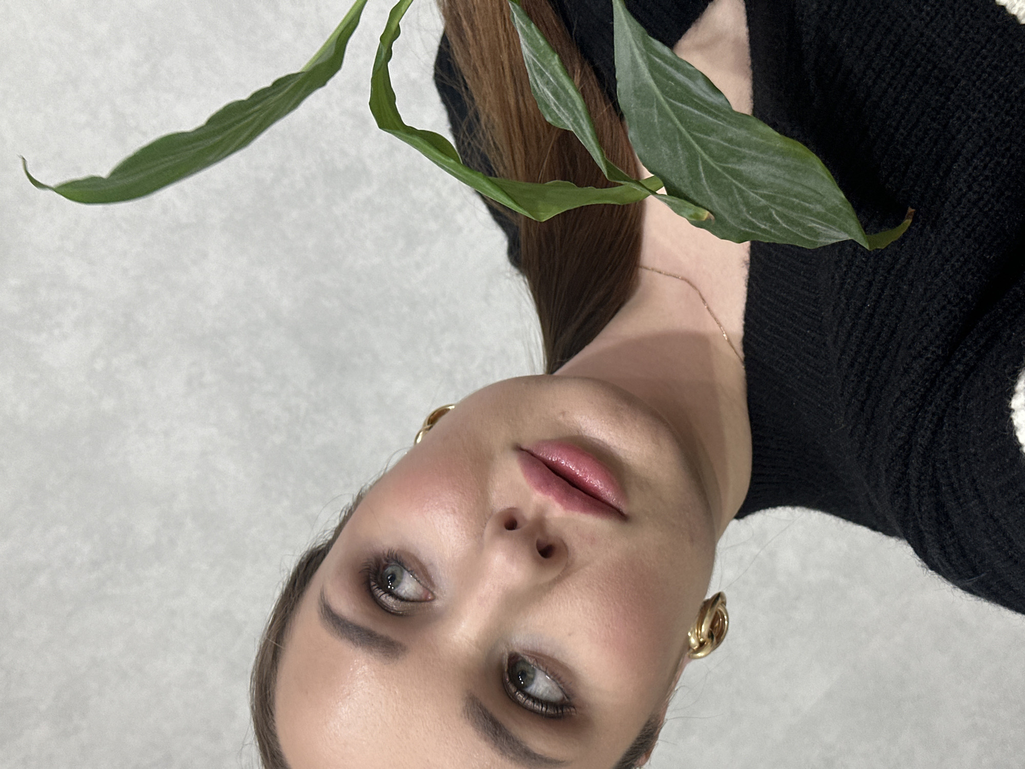 A woman with light skin, brown hair pulled back in a ponytail, and wearing gold hoop earrings is pictured from a slightly low angle. She is facing the camera with her head tilted to the right. She has light eye makeup and her lips are glossy. Part of a green plant with several leaves is visible in the upper left and upper right, obscuring portions of her face and background. She is wearing a black sweater. The background is a light gray color. There is no indication of the location being Chaikovsky, Russia.