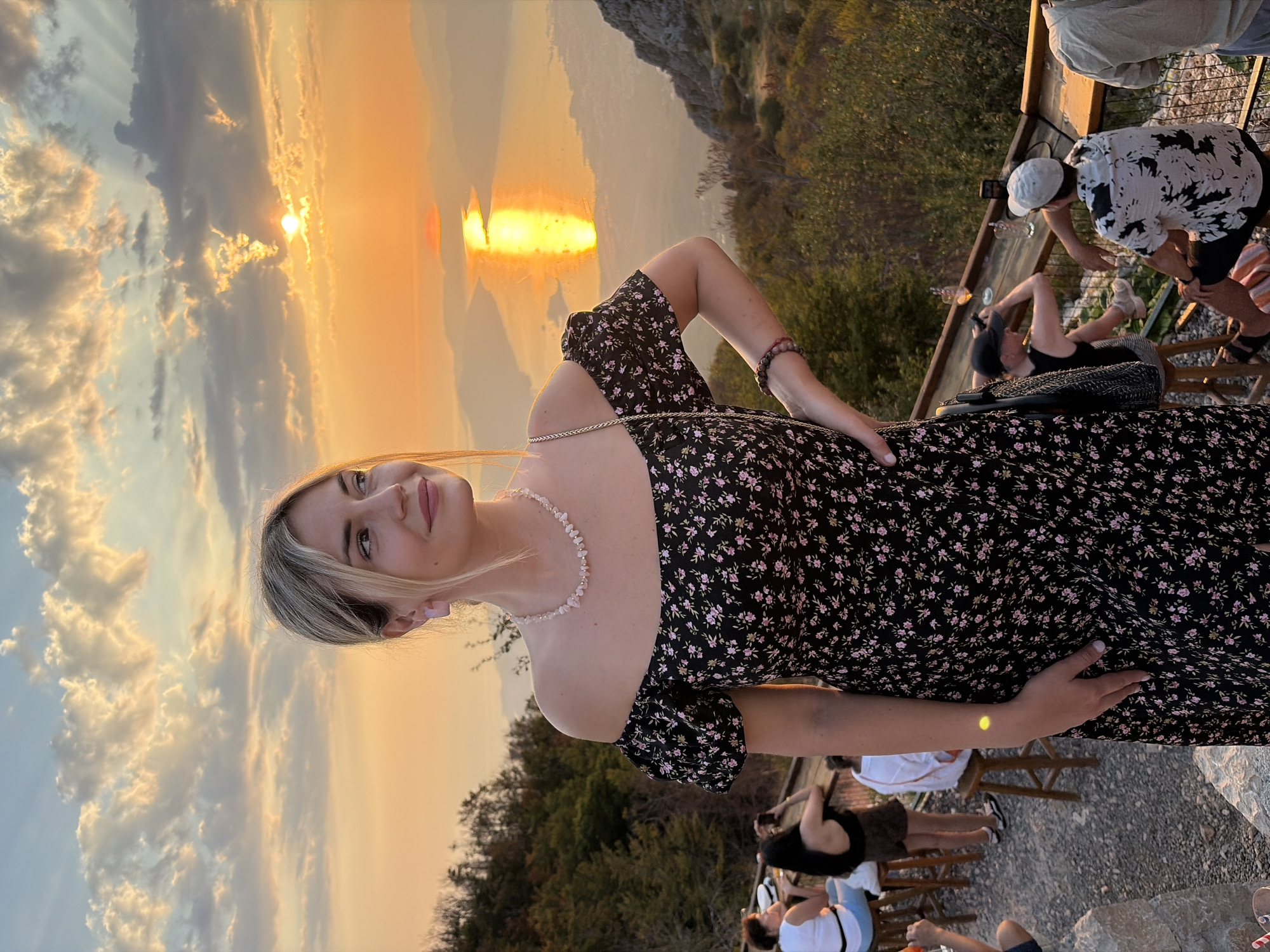 In the city of Kuk, Montenegro, a young woman with blonde hair and fair skin, wearing an off-the-shoulder floral dress and a beaded necklace, poses for a photograph against a backdrop of a dramatic sunset over a body of water. The sun, a bright orange orb, is partially obscured by clouds, casting a golden reflection on the water below and illuminating the surrounding mountainous landscape. In the foreground, other individuals are seated at tables, enjoying drinks and the view. One woman is taking a photograph, while others are engaged in conversation. The overall scene depicts a social gathering in a scenic, elevated location, capturing the beauty of a sunset.
