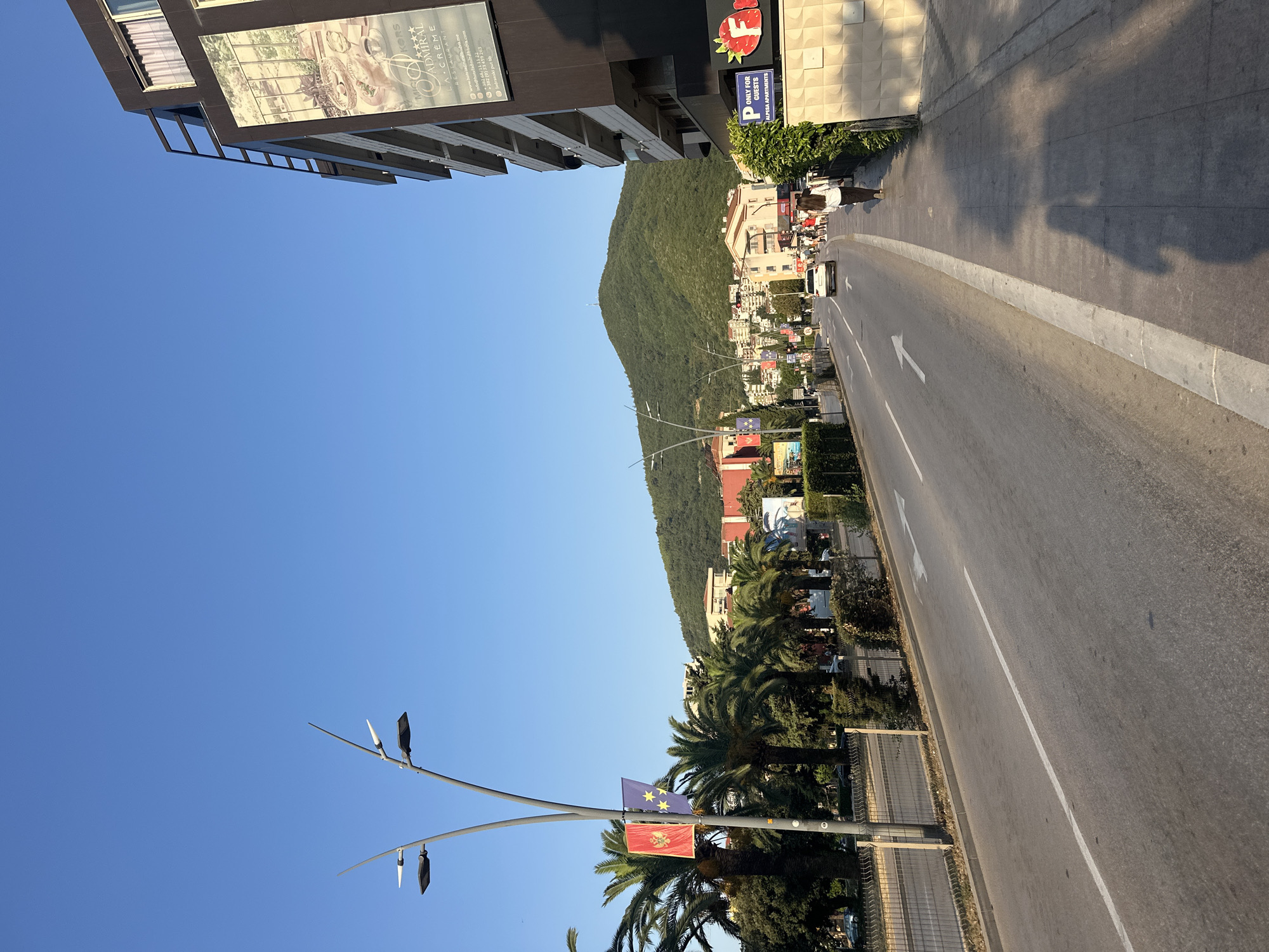 The image depicts a street scene in Budva, Montenegro, on a clear, sunny day. A paved road with white lane markings runs diagonally from the bottom left to the upper right of the frame. On the right side of the road, a sidewalk is visible, with shadows of people cast upon it. A woman is walking away from the camera on the sidewalk. Adjacent to the sidewalk is a modern building with dark facade and balconies, and a large banner advertising a hotel or restaurant. On the left side of the road, there are palm trees and vegetation, a metal fence, and streetlights with modern designs. In the background, a lush green hill dominates the skyline, with buildings clustered on its slopes and at its base. The sky is a clear, vibrant blue.