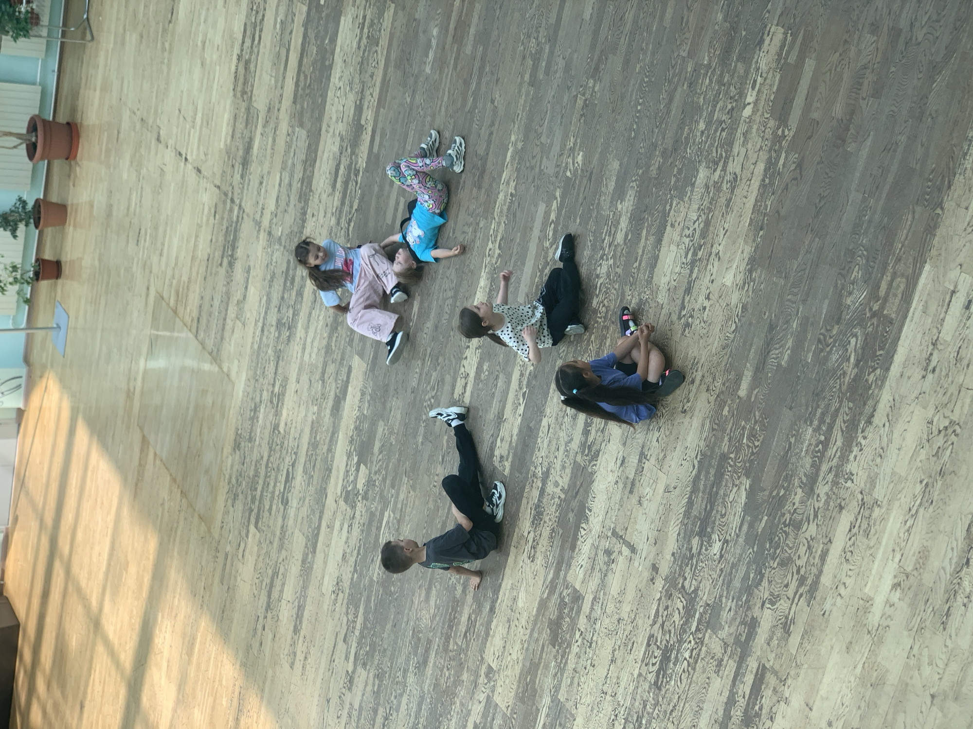 Six children are seated on a light brown wooden floor, with sunlight casting diagonal shadows from the left. The children are in various resting positions. In the upper portion of the image, a girl in light blue sweatpants and a dark t-shirt sits cross-legged, looking down. To her right and slightly below, another girl in a blue t-shirt and brightly patterned leggings lies on her back, looking upwards. To the left and slightly lower, a boy in a dark grey t-shirt and black pants sits with his legs extended to the right, looking to the right. Below him and slightly to the right, a girl with dark hair in a ponytail wears a white t-shirt with black polka dots and black pants, looking to her left. Further down and to the left, a girl with dark hair in a ponytail wears a blue t-shirt and dark shorts, looking down. Another child, partially visible at the bottom edge, wears dark shorts and what appears to be a blue t-shirt, looking down. The environment appears to be an indoor space, possibly a gymnasium or large hall, with large windows or skylights allowing for significant natural light. The wooden floor has a distressed or aged appearance with visible grain and variations in color. The scene takes place in Megion, Russia.