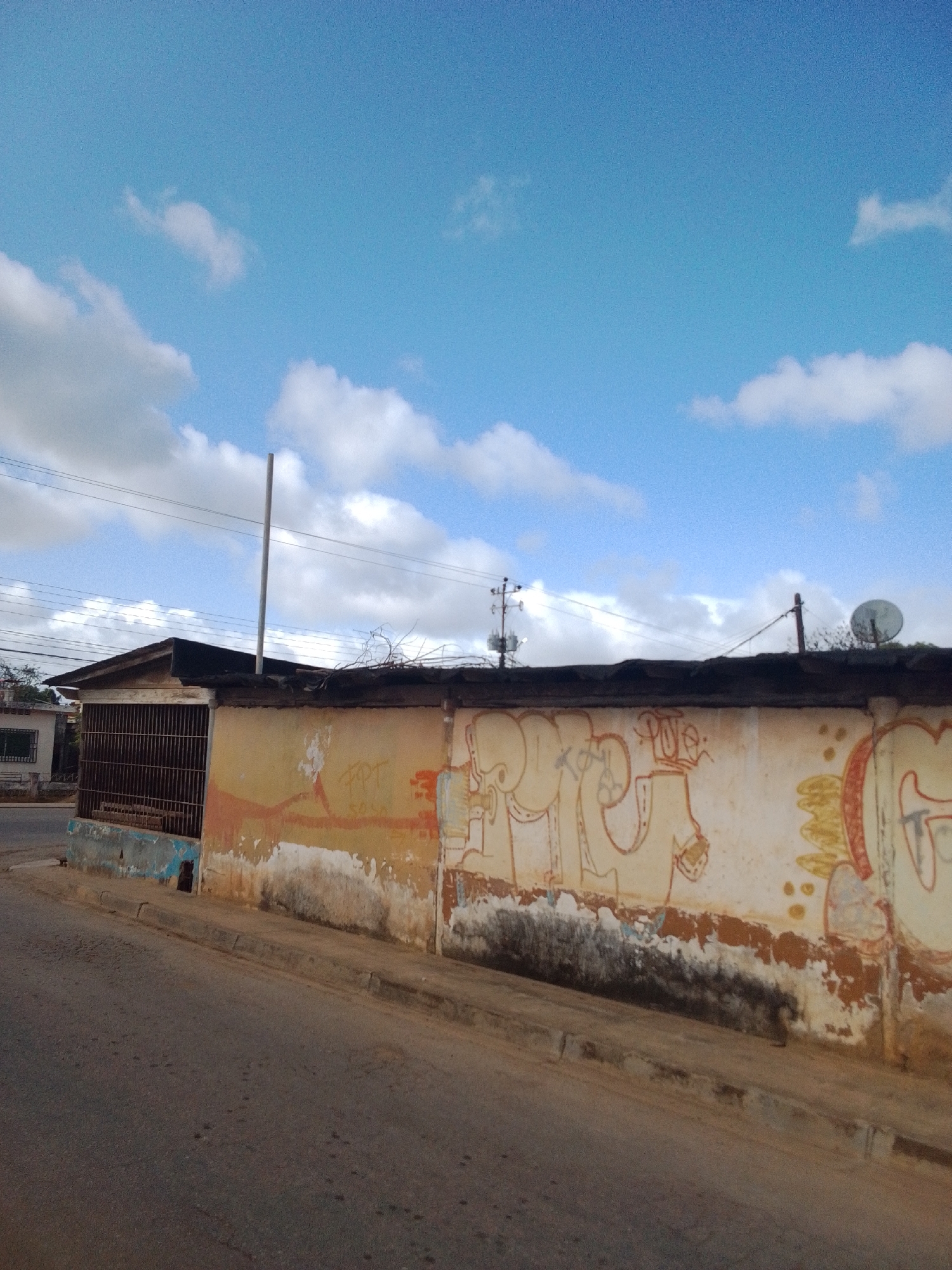 The image depicts a street scene in Altagracia, Venezuela. A weathered, tan-colored wall with graffiti runs horizontally across the frame, partially obscuring a building behind it. The graffiti features abstract figures and lettering in red, orange, and yellow. To the left of the wall, a building with a dark-sloped roof and a barred window is visible. A tall, thin pole extends vertically from the roof of this building. Overhead, a bright blue sky is dotted with white clouds. Electrical wires are strung across the scene, supported by utility poles. A satellite dish is mounted on a pole on the right side of the frame, above the wall. The foreground shows a gray asphalt road and a concrete curb.