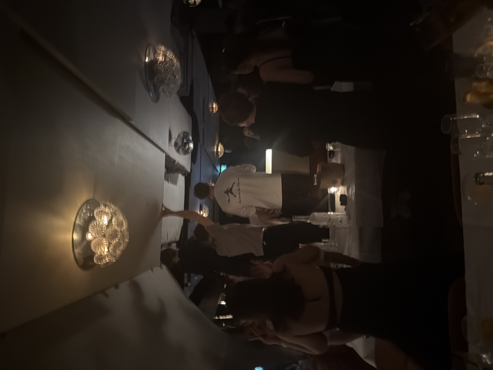 The image shows a dimly lit indoor space with people standing around tables. There are light fixtures on the wall. Some people are wearing casual clothes, and there appear to be drinks on the tables. The location is unknown.
