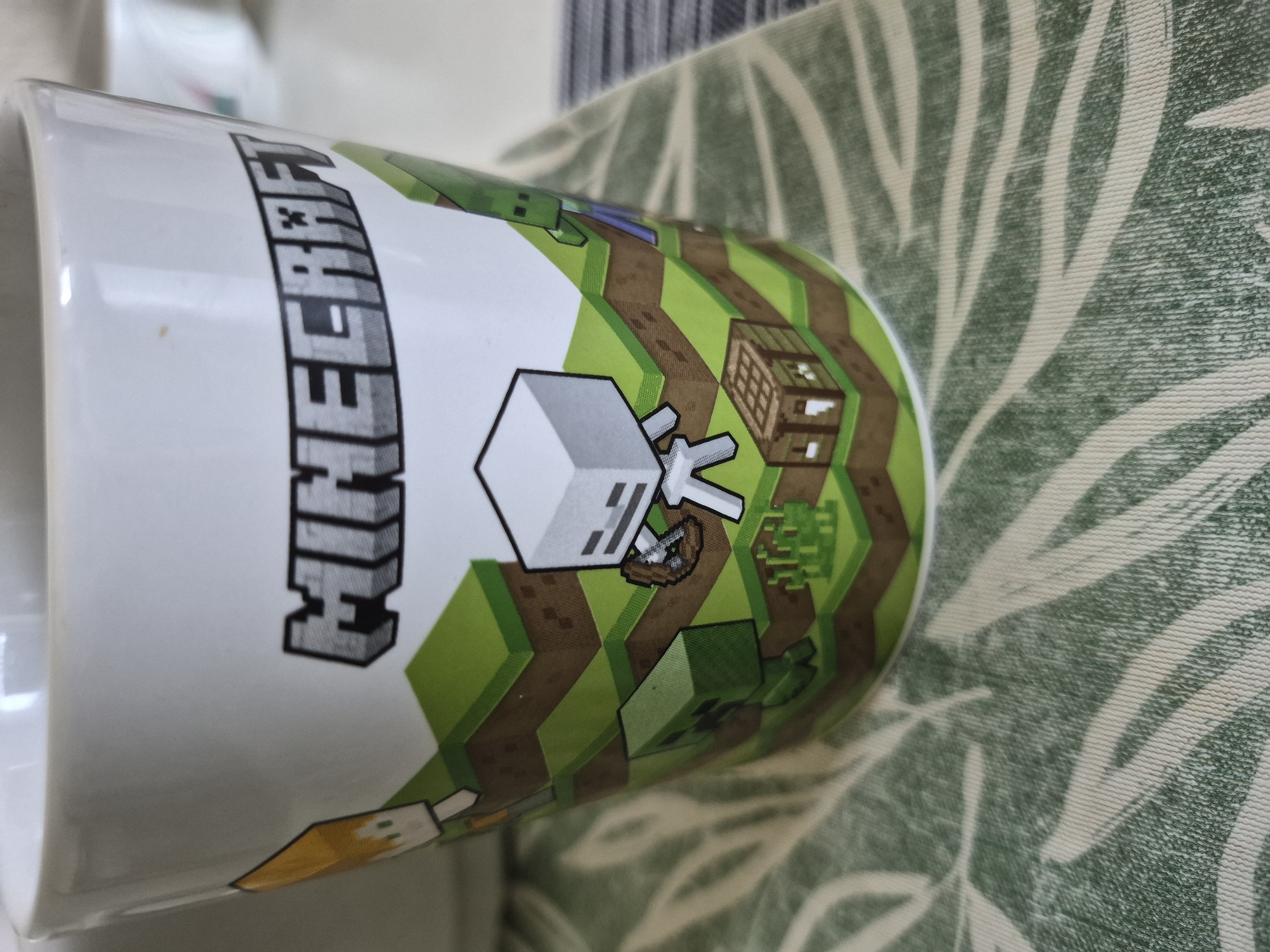 The image is a close-up shot of a white ceramic mug with a Minecraft-themed design. The design features pixelated elements characteristic of the game, including a character resembling Steve with orange hair and a blue shirt, a skeleton character holding a bow, a green Creeper, and a small wooden crafting table. The background of the design depicts a layered landscape of green grass and brown dirt blocks. The mug is placed on a green and white patterned placemat. The overall lighting suggests an indoor setting, possibly on a table. The location is Unknown, Unknown.