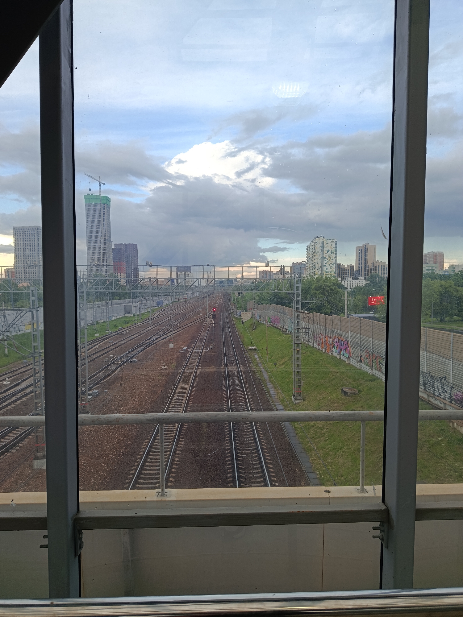 A ground-level, medium-shot captures a multi-track railway system, likely associated with a high-speed transit or commuter rail line, extending into the distance. The view is framed by a window or glass enclosure, suggesting the image was taken from a bridge, overpass, or similar elevated structure. The train tracks are rusty-brown in color.
Several high-rise buildings are visible in the distance, scattered along the cityscape. The sky is overcast with grey clouds.
The location is Moscow, Russia.