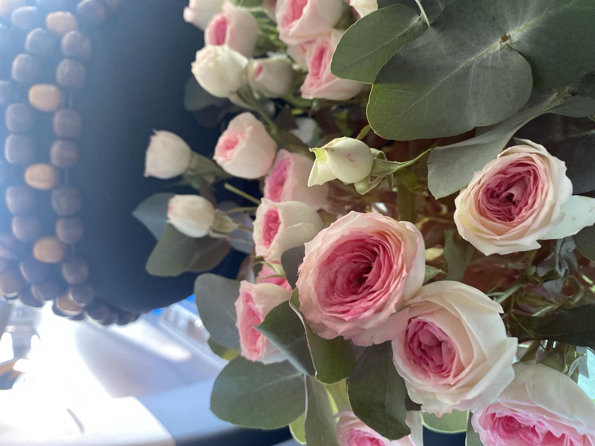 The image depicts a close-up of a bouquet of pink and white roses with eucalyptus leaves. The flowers are arranged in a way that suggests they are part of a larger display or arrangement, possibly indoors. The background is blurred, but hints of a dark, textured object, possibly a vase or decorative element, can be seen. The lighting suggests an indoor setting, with some ambient light sources creating highlights and shadows on the petals and leaves. There is no visible indication of the city of Pavlodar, Kazakhstan, or any specific location within it. The image focuses solely on the floral arrangement.