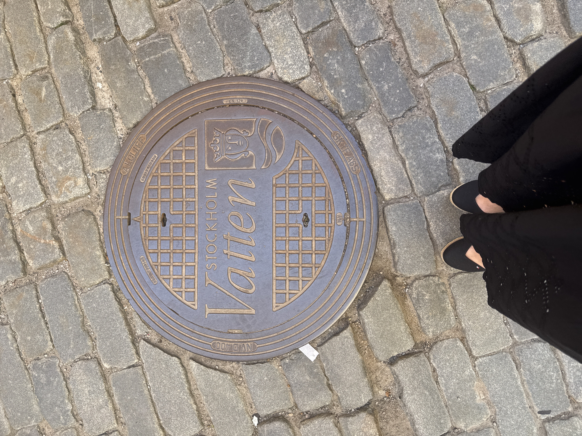 A person's feet, wearing black flats and a black dress, are visible in the foreground, standing on a cobblestone street. In the center of the image is a circular manhole cover with the inscription "STOCKHOLM VATTEN" and a shield with a crown and a face. The manhole cover has additional inscriptions around its edge, including "CALCLIFE", "NVD 400", "EN 124", and "SKIFAB69". The environment is an outdoor, urban setting with cobblestone paving.