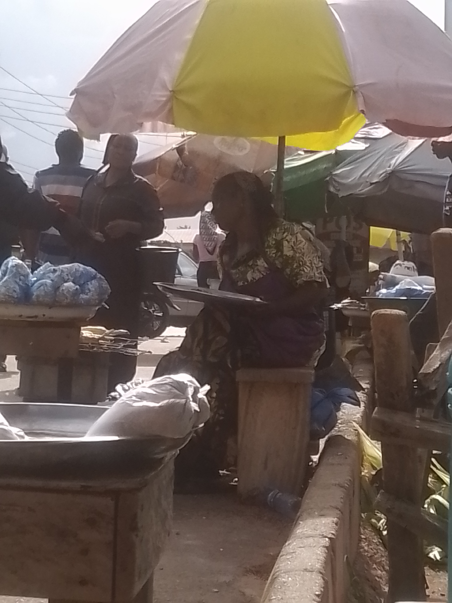 An outdoor marketplace scene is depicted, with a woman seated on a stool, holding a document or book. She is wearing a patterned top and a purple skirt. Behind her, a large yellow and white umbrella provides shade. Several other people are visible in the background, some appearing to be vendors. There are market stalls with various goods, including blue-colored items on display in a bin to the left. In the foreground, a large metal basin or container is partially visible. The ground appears to be dirt or gravel. The overall atmosphere suggests a bustling, commercial environment.