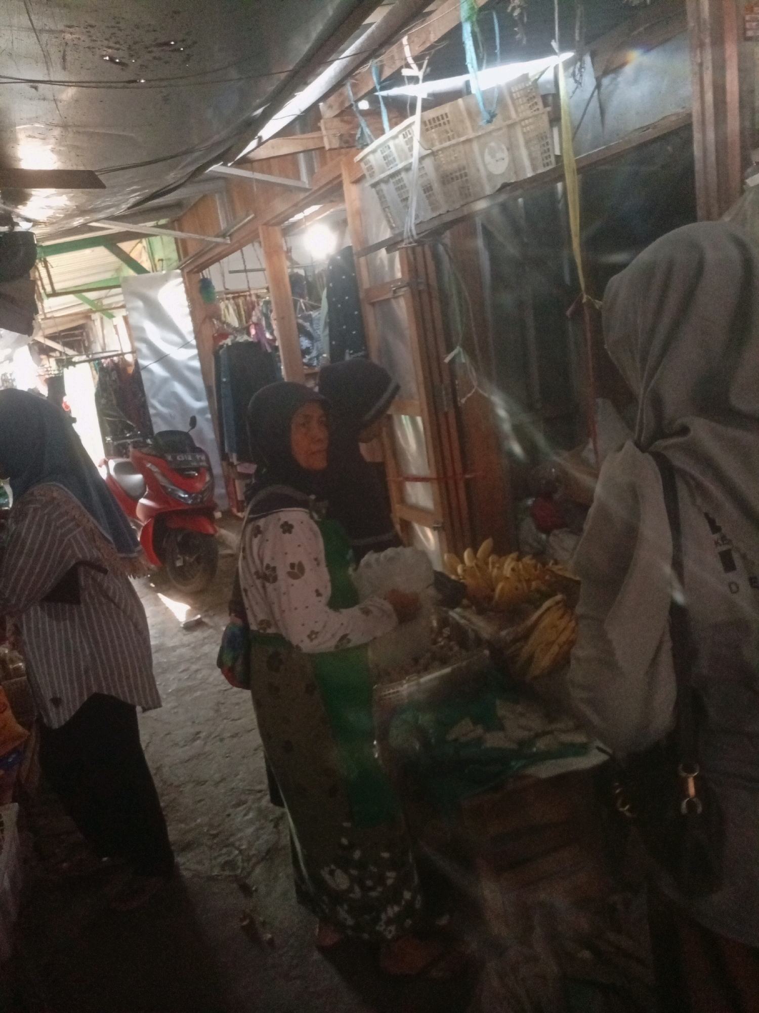 The image depicts a street market scene in Trahan, Indonesia. The market appears to be an open-air setup with stalls constructed from wood and covered with tarps. In the foreground, to the left, a woman in a striped long-sleeved shirt and dark pants is partially visible. To her right, a woman wearing a hijab and a long patterned skirt is standing, holding a white object. Further to the right, another person, also wearing a hijab, is seen from behind, their head and shoulders draped in a grey garment. In the mid-ground, a red scooter is parked. Various goods are displayed, including what appear to be bunches of bananas on a stall. The environment is dimly lit, suggesting it could be early morning or late afternoon, or simply a shaded area within the market. The overall impression is that of a bustling local marketplace.