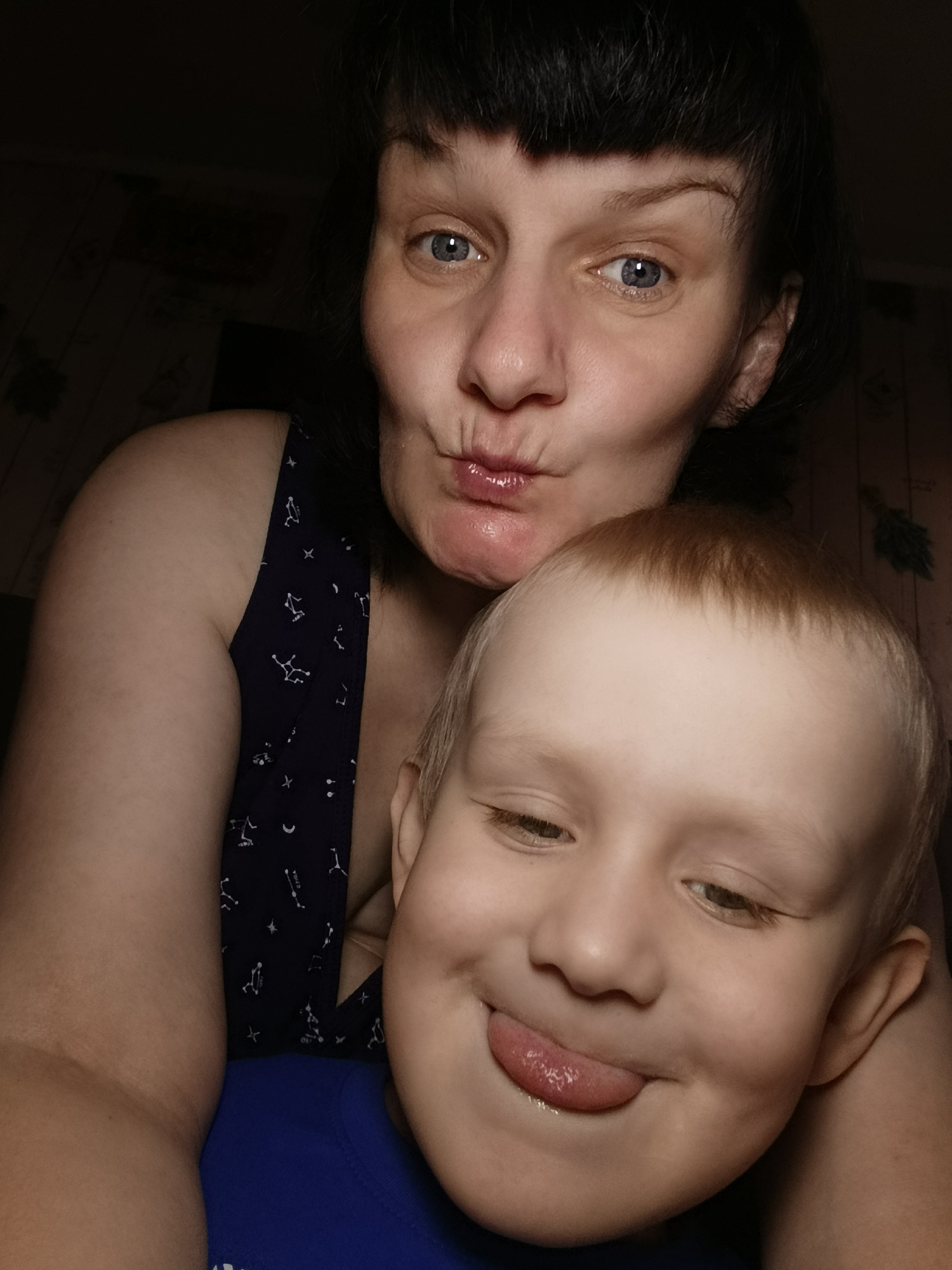 A close-up photo captures a woman and a young boy. The woman is at the top of the frame, puckering her lips for a kiss, with blue eyes and dark, short-cropped hair. She wears a dark tank top with a pattern of small white constellations. Beneath her, a blond boy is sticking out his tongue, his eyes half-closed in a playful expression. He is wearing a blue shirt. The background is dark, suggesting an indoor setting with low lighting.