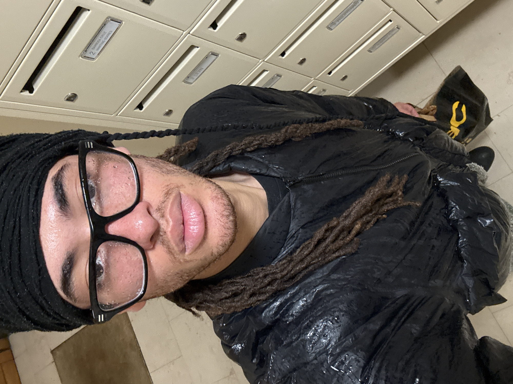 A person with dreadlocks wearing glasses, a black head covering, and a black jacket is in what appears to be an indoor hallway with mailboxes. The person is close to the camera, and the mailboxes are in the background. To the right of the person is a black shopping bag with a yellow logo. The location cannot be determined to be Paris, France based on the image provided.