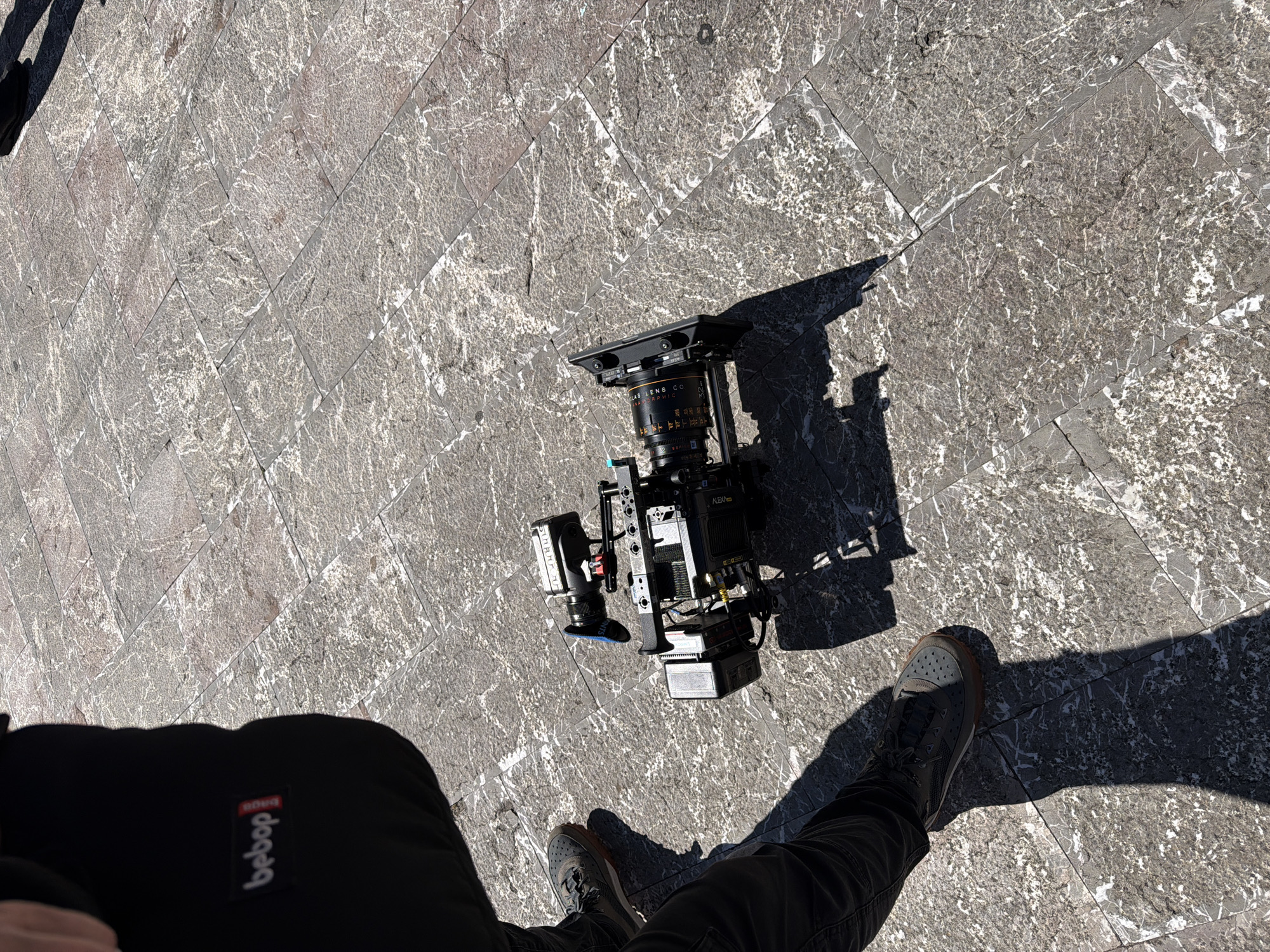 A high-angle shot depicts a person standing on a stone-tiled surface alongside a professional film camera. The camera is black with an attached lens, monitoring screen, and other accessories. The person is wearing dark pants with a "bebob" logo visible on the left leg and dark athletic shoes. The stone tiles are grey with a marbled pattern. The camera's shadow is cast towards the right, and the person's shadow also extends onto the stone tiles. Based on the scene's appearance, the photograph was taken in Itala, Italy.
