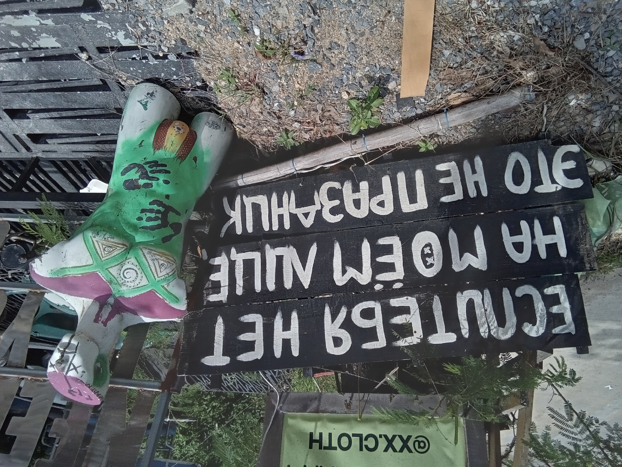 In Koh Phangan, Thailand, a dark wooden sign with white lettering reads "ЕСЛИ ТЕБЯ НЕТ НА МОЁМ ЛИЦЕ - ЭТО НЕ ПРАЗАНЧИК". To the right of the sign, a headless, partially painted mannequin is positioned. The mannequin is painted green with purple accents on the chest and head. It also features intricate green and white patterns on its torso. The scene is set outdoors, with gravelly ground and some sparse vegetation visible. A pale green banner with black text "@XX.CLOTH" is visible behind the sign.