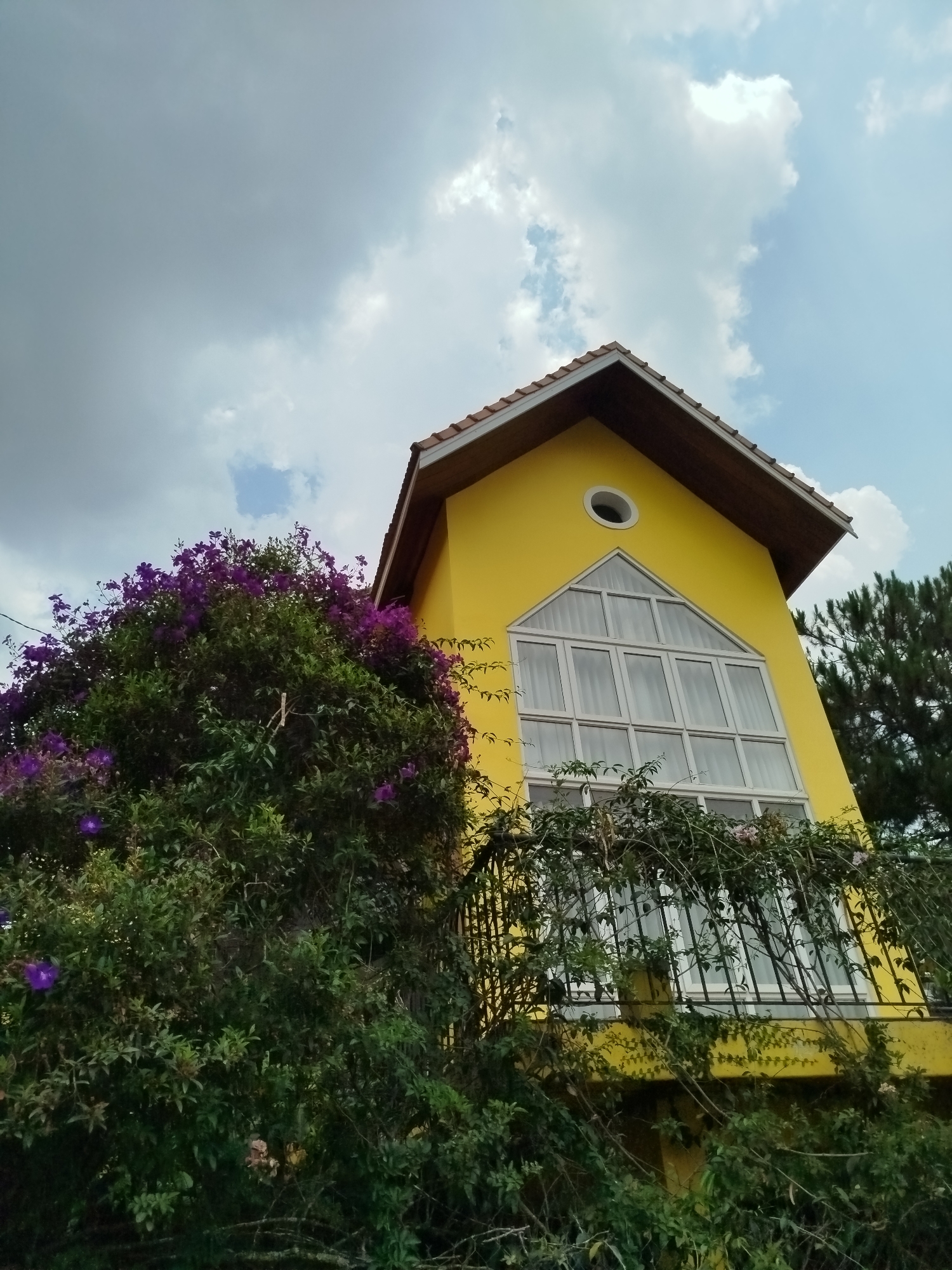 A low-angle shot depicts a bright yellow house with a gable roof located in Hiep Thanh, Vietnam. The house features a large, triangular window divided into multiple panes and a smaller, round window above it. A wrought-iron balcony partially obscures the lower portion of the window, with green vines and foliage growing over and around it. To the left of the house, a large bush with purple flowers is prominently displayed. In the background, a cloudy sky with patches of blue is visible. A tall pine tree is present on the right side of the house.