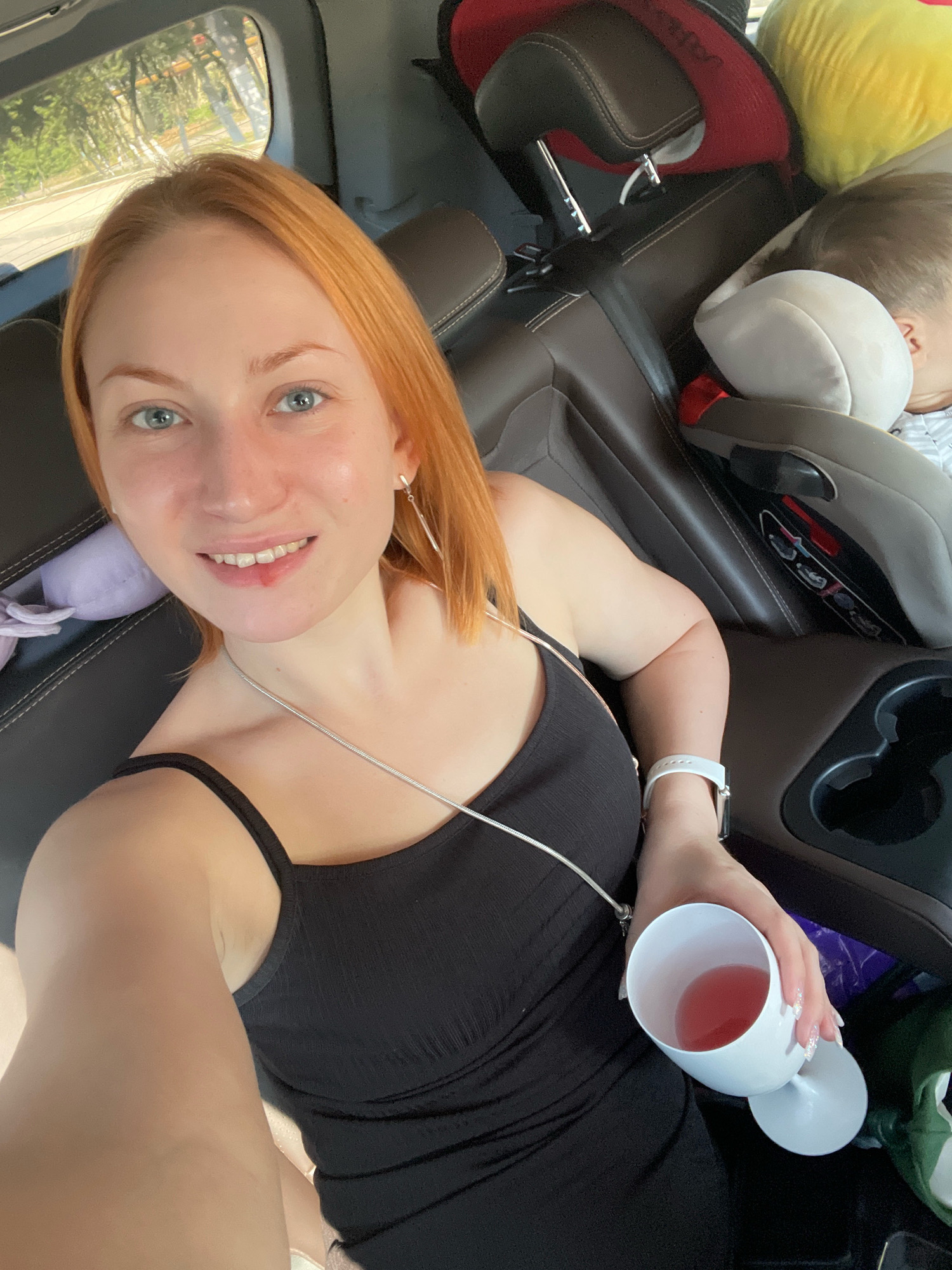 A woman with auburn hair and blue eyes is taking a selfie inside a vehicle. She is wearing a black tank top and a silver necklace. She is holding a white wine glass filled with a pink beverage. A child's car seat is visible in the background, and a child is sleeping in it. The woman has a small red mark on her upper lip. The interior of the vehicle appears to be made of dark leather. The scene is likely in Shkolny, Russia, given the context, but this cannot be definitively determined from the image alone.