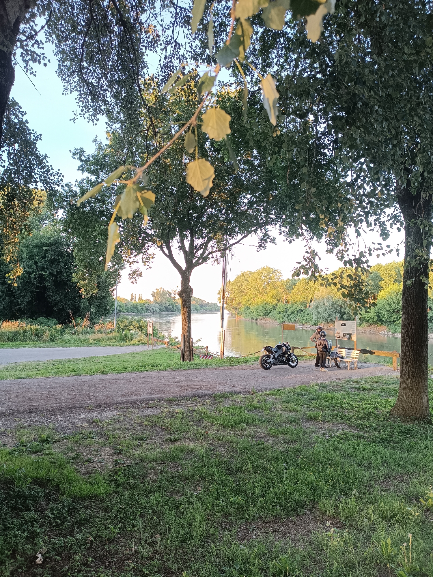 The image depicts a riverside scene in Marcaria, Italy, at what appears to be late afternoon or early evening. The foreground is dominated by grass and a gravel path leading to a paved road that runs parallel to the river. Large trees with dense foliage provide shade, with some leaves in the foreground obscuring the view.
In the middle ground, beside the gravel path, a black motorcycle is parked. Two individuals, a man and a woman, are standing near the motorcycle. Both are dressed in motorcycle gear, including helmets, suggesting they have either just arrived or are preparing to depart. They are facing each other and appear to be engaged in conversation or a brief interaction. A wooden bench is situated near them.
The river itself is a prominent feature, flowing calmly with its surface reflecting the warm, golden light of the setting sun. The opposite bank is lined with lush green trees and vegetation, creating a natural and tranquil backdrop. In the distance, more trees and a clear sky are visible. There are also some signs and a wooden barrier near the riverbank. The overall atmosphere is peaceful and serene, capturing a moment of pause during a motorcycle journey.