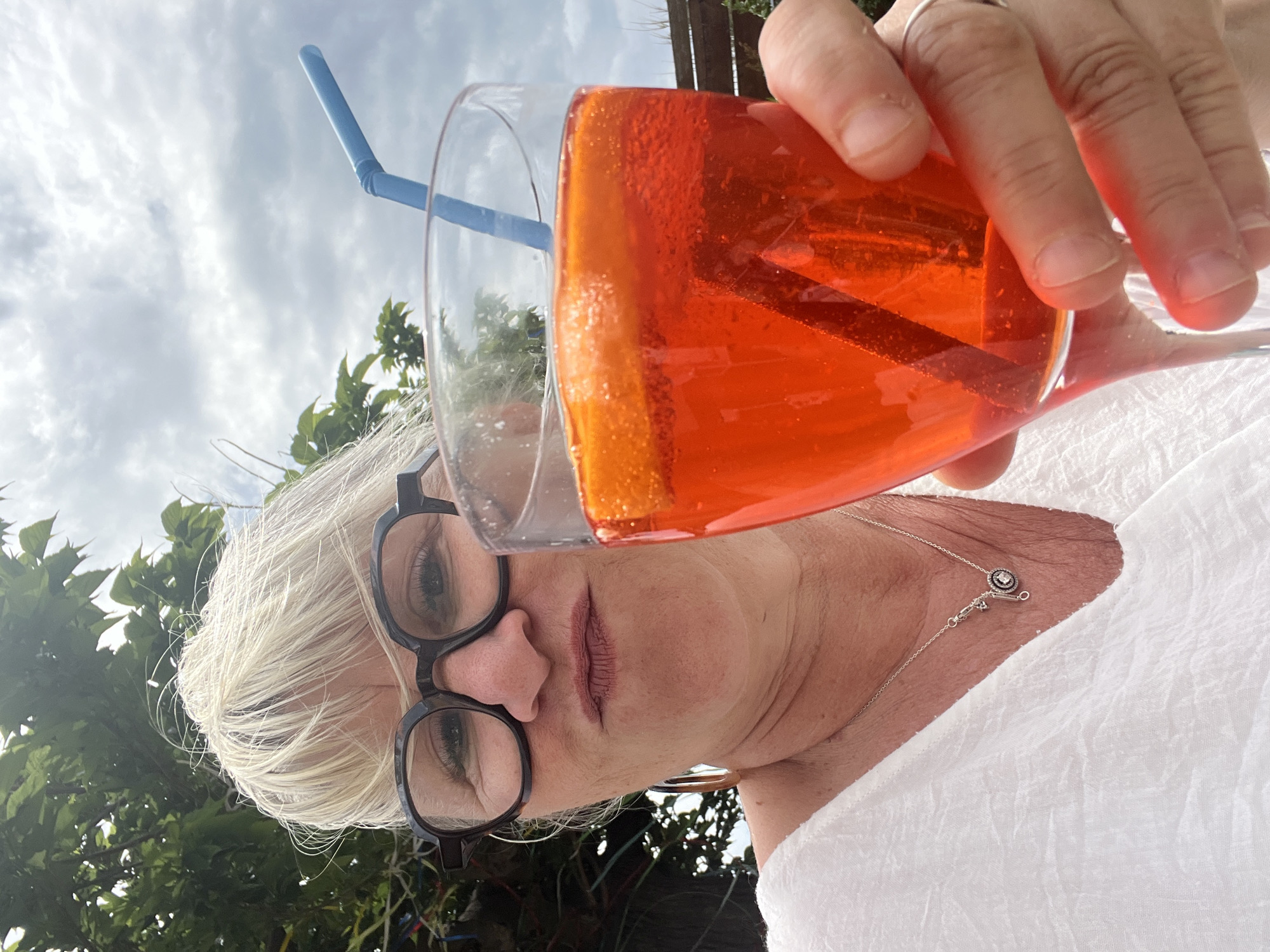 A middle-aged woman with short, white hair and glasses is holding a glass of orange liquid with a blue straw. The drink is in the foreground, obscuring part of her face. She is wearing a white shirt and a silver necklace with a circular pendant. The background shows a sky with scattered clouds and some green foliage, suggesting an outdoor setting. The location is Montarnaud, France, though no specific landmarks are visible.