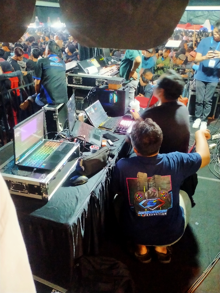 The image shows a scene from an event held in Boyolali, Indonesia, at the coordinates -7.535 latitude, 110.601 longitude. The image shows a partially obscured view of a stage, with several people sitting in front of a table with a variety of electronics. The electronic items are arranged in a seemingly organized manner. The equipment includes four laptops, a keyboard, a computer monitor, and a small speaker. The table has a black cloth draped over it, and there is a black chair in the foreground. The scene is partially lit, with the shadows appearing behind the stage. The people in the image are wearing casual clothing and appear to be focused on their tasks. The environment is an outdoor area, possibly an event space or concert hall. The view is from the edge of the stage, behind the people. The image is captured from a slightly elevated position.