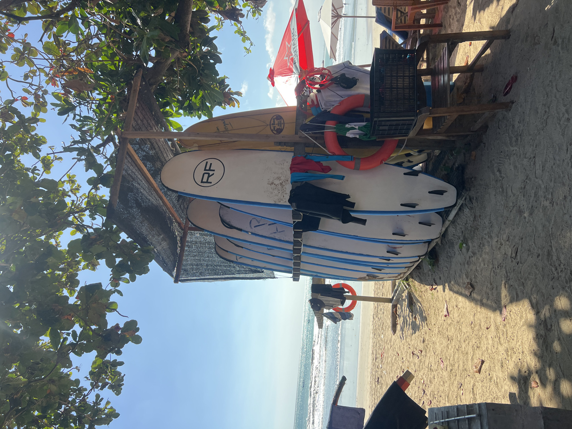 The image depicts a beach scene in Kuta, Indonesia, likely Kuta Beach, known for its surf spots. In the foreground, a rack holds several surfboards upright, some with blue accents and one with "RF" branding. Attached to the rack are various items of clothing, including wetsuits, and two orange lifebuoys. To the right, a wooden chair supports a black crate, and beyond that, beach chairs and a red umbrella are visible. The sandy beach extends to the ocean, where waves are breaking under a clear blue sky. Lush green trees with dappled sunlight cast shadows on the sand.