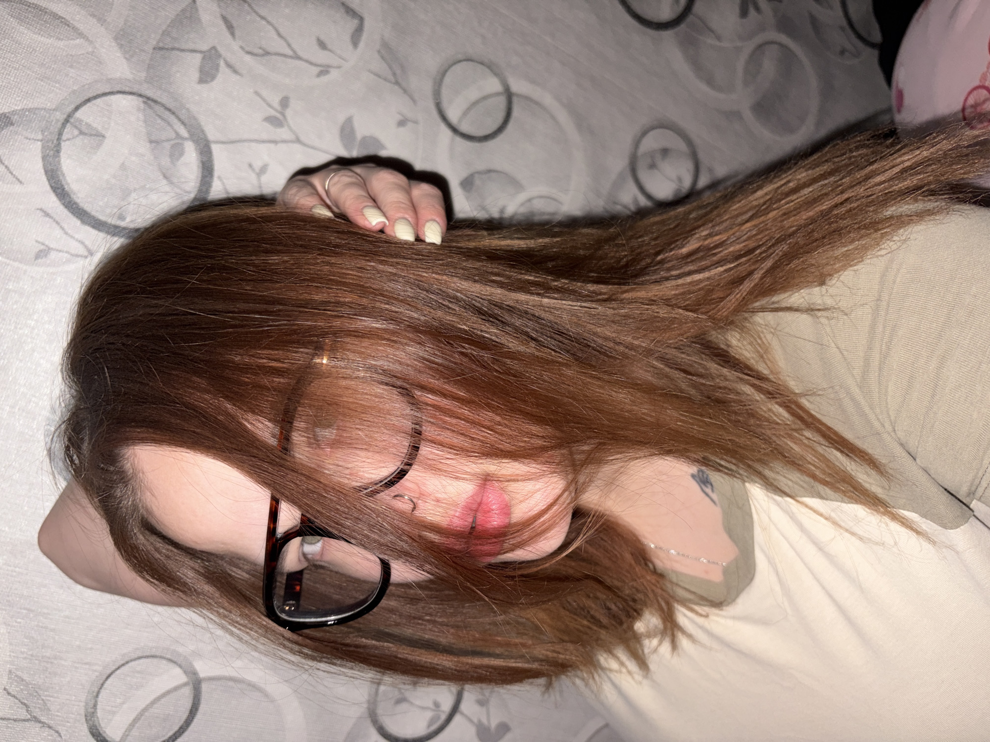 A woman with long, auburn hair is lying down. She wears glasses with a dark frame. Her left hand is visible, resting on her hair near her head. She has a lip piercing. A silver necklace is around her neck. She is wearing a light beige shirt. The background shows a patterned surface, likely a bed or couch covering, with large gray circles. There is no evidence to indicate this photo was taken in Novomargaritovo, Russia.