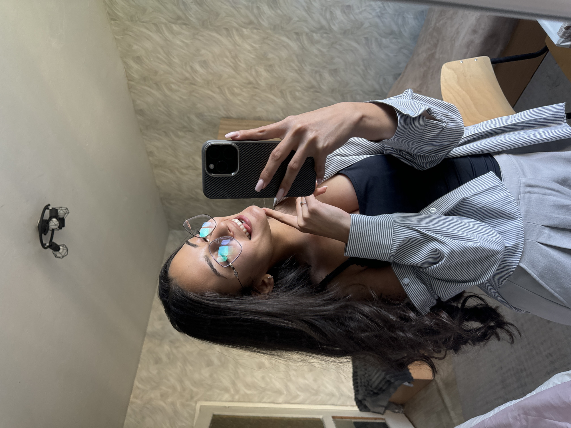 A woman is taking a selfie indoors. She is wearing glasses, a black top, and a striped shirt. She is holding a phone with a black case. The background includes a textured wall, a light fixture, and a glimpse of a wooden chair. The location in the city of Sofia, Bulgaria is unknown.