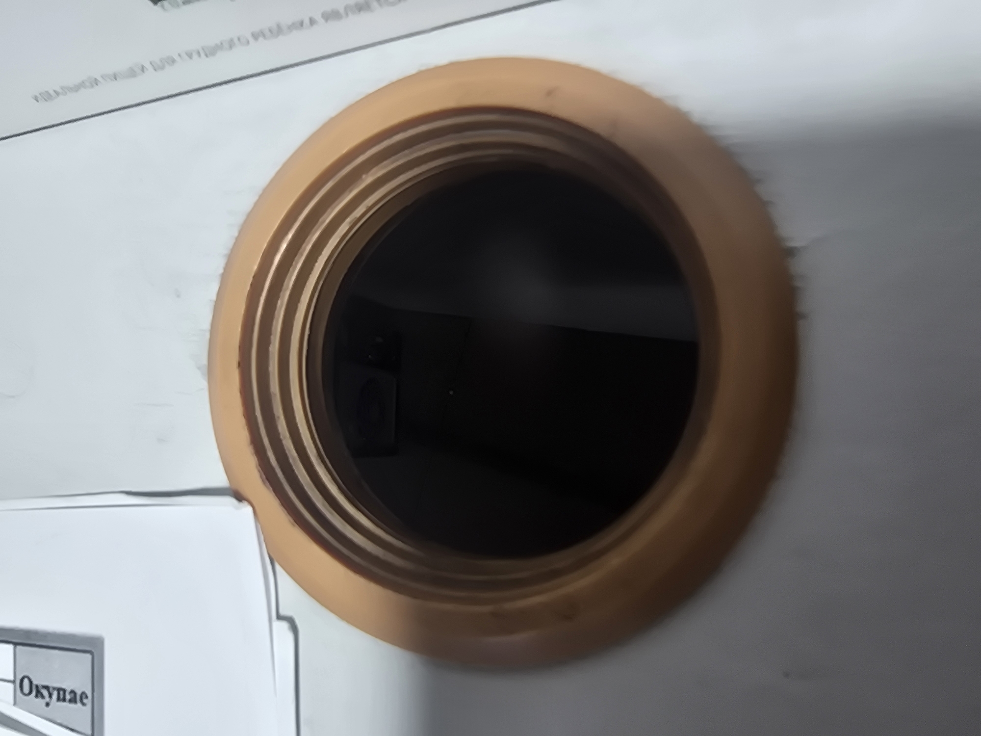 A top-down, medium-shot photograph captures a close-up of a beige, threaded plastic rim surrounding a dark, circular opening. The opening appears to be a hole or a receptacle, the depth and contents of which are not fully discernible due to the darkness. The rim has a ribbed texture and appears to be made of a smooth, molded material.
To the left of the rim, a white sheet of paper is partially visible. On this paper, a dark rectangular box encloses text written in an upright, sans-serif font. The visible portion of the text reads "Oxyone". The paper itself has a slight sheen and appears to be a standard document.
To the right of the rim, another white document is present, its surface partially covered by a thin, black line that runs diagonally from the top right corner towards the center. Faint, smaller text is visible along this line, written in a small, sans-serif font, but it is illegible.
The overall environment appears to be an indoor setting, likely a desk or table surface, given the presence of documents and the object's placement. There are no individuals visible in the image, nor any actions or interactions taking place. The lighting is even, casting no harsh shadows and highlighting the textures of the plastic rim and the paper. The background is a plain, light-colored surface, possibly a wall or table, with some subtle smudges or imperfections. The image is focused on the rim and opening, with the surrounding elements slightly out of focus.
This scene is not specifically identifiable as being located in the city of Ulan-Ude, Russia. The provided information does not contain any elements that directly indicate this geographical location.