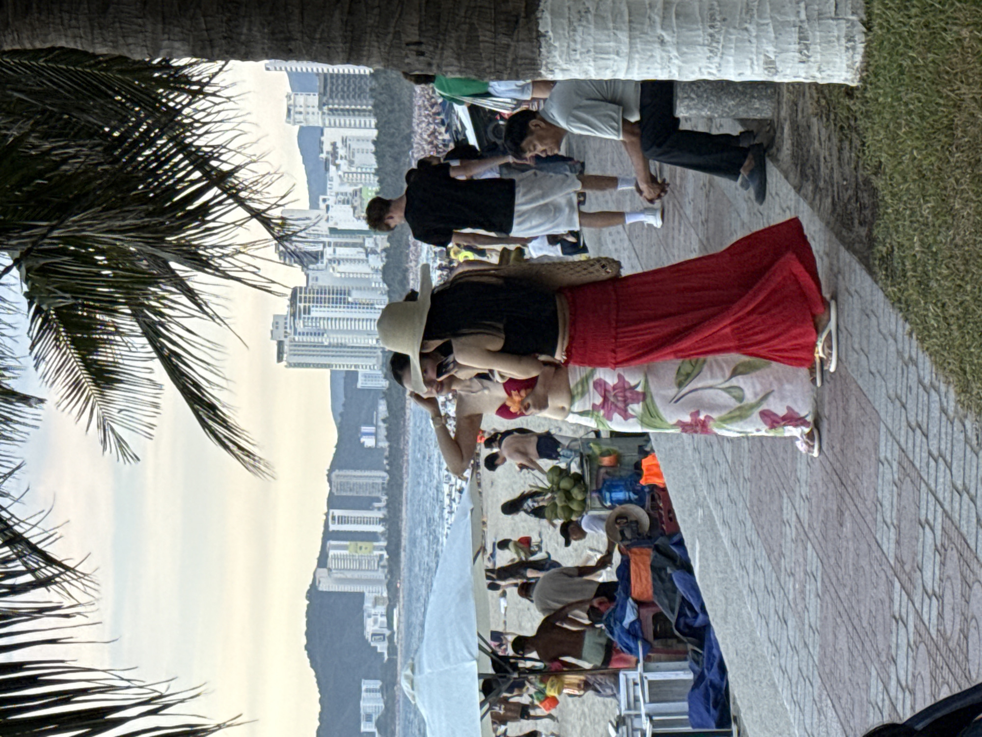 The image shows a group of people on a paved walkway next to a grassy area and a beach. In the background, there is a city skyline with many tall buildings and a body of water. Palm fronds are visible in the foreground, framing the scene. The people are dressed in casual summer clothing, suggesting warm weather. Some are standing, while others appear to be sitting or walking. One person in the foreground is wearing a wide-brimmed hat and a red skirt, and another is wearing a floral-patterned skirt. The atmosphere seems relaxed and leisurely, possibly indicating a vacation or a day at the beach. The time of day appears to be late afternoon or early evening, given the soft lighting. The presence of the city and beach suggests a coastal urban setting, consistent with Nha Trang, Vietnam. No text is visible in the image.