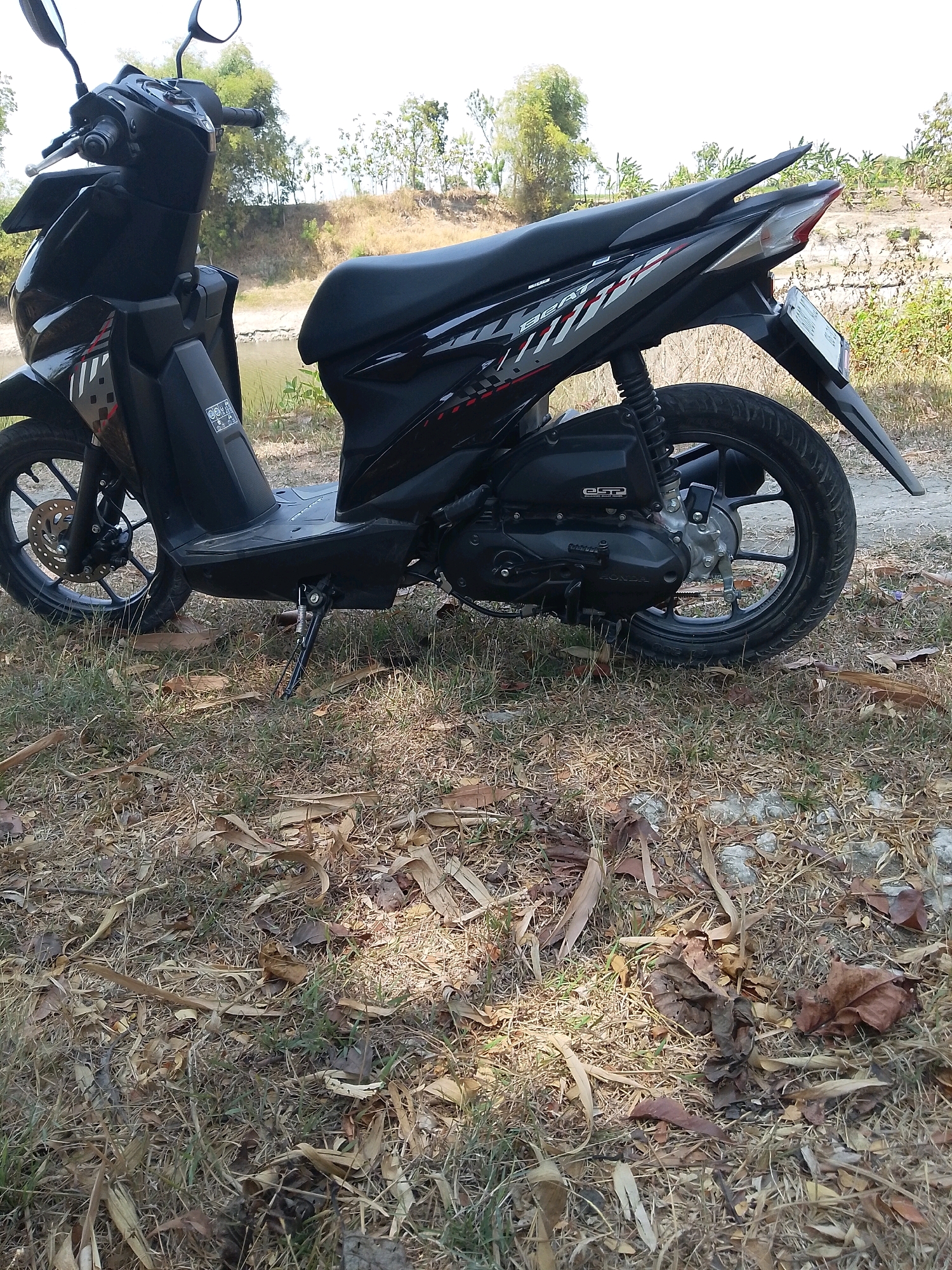 A black Honda Beat scooter is parked upright on its kickstand on dry, patchy grass in Grebegan, Indonesia. The scooter is positioned at an angle, with the front wheel facing left and the rear wheel facing right. The scooter's seat is black, and it has a black body with red and silver decals. The handlebars and mirrors are visible at the top left of the image. In the background, there is a body of water, lush green vegetation, and a slightly elevated dirt path. The sky is bright and overcast.