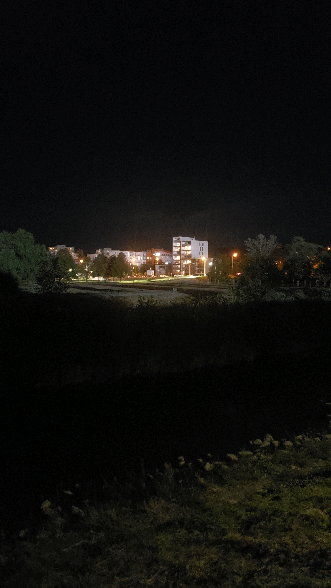 The image shows a nighttime view of a residential area in Prešov, Slovakia. In the background, several apartment buildings are illuminated by interior lights and streetlights. A prominent white building with multiple floors and windows is visible on the right side. Trees are scattered throughout the scene, some of which are silhouetted against the lit buildings. In the foreground, there is a dark, grassy embankment with scattered rocks. The overall atmosphere is calm and quiet, typical of a residential neighborhood at night.