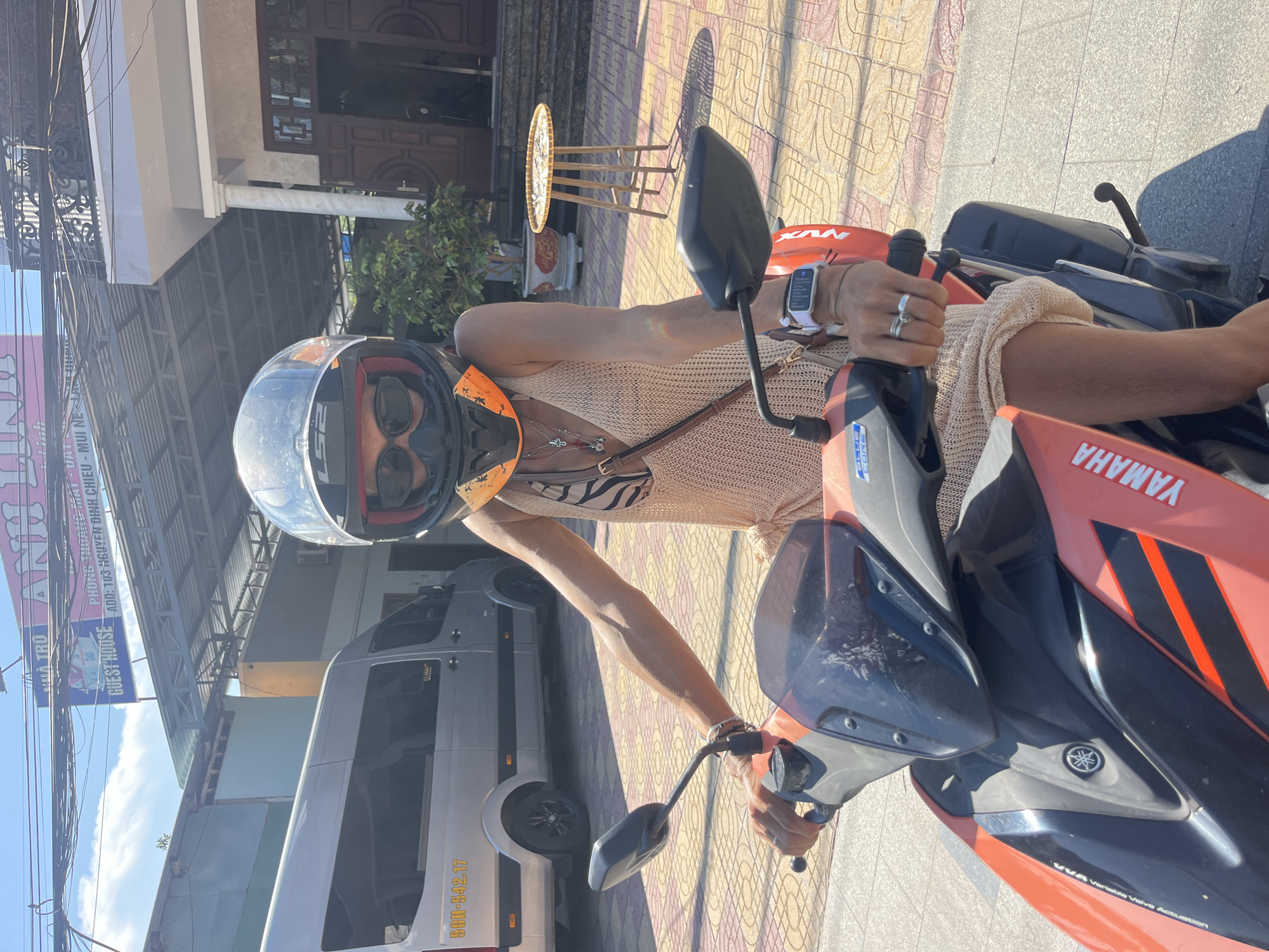 A woman wearing a helmet and sunglasses is sitting on an orange and black Yamaha scooter. She is wearing a light beige knitted dress and a striped bikini top underneath. She has rings on her fingers and a smartwatch on her left wrist. The scooter is parked on a paved sidewalk with a patterned tile design. In the background, a silver van is parked, and a building with a blue and pink sign is visible. The sign reads "NHÀ TRÚ ANH LINH GUEST HOUSE PHÒNG TRỌ CỰC CHẤT ADD: 103 NGUYỄN ĐÌNH CHIỂU - MŨI NÉ - PHAN THIẾT." This indicates the location is a guest house on Nguyen Dinh Chieu street in Mui Ne, Phan Thiet, Vietnam. The environment appears to be a sunny day in a residential or tourist area.