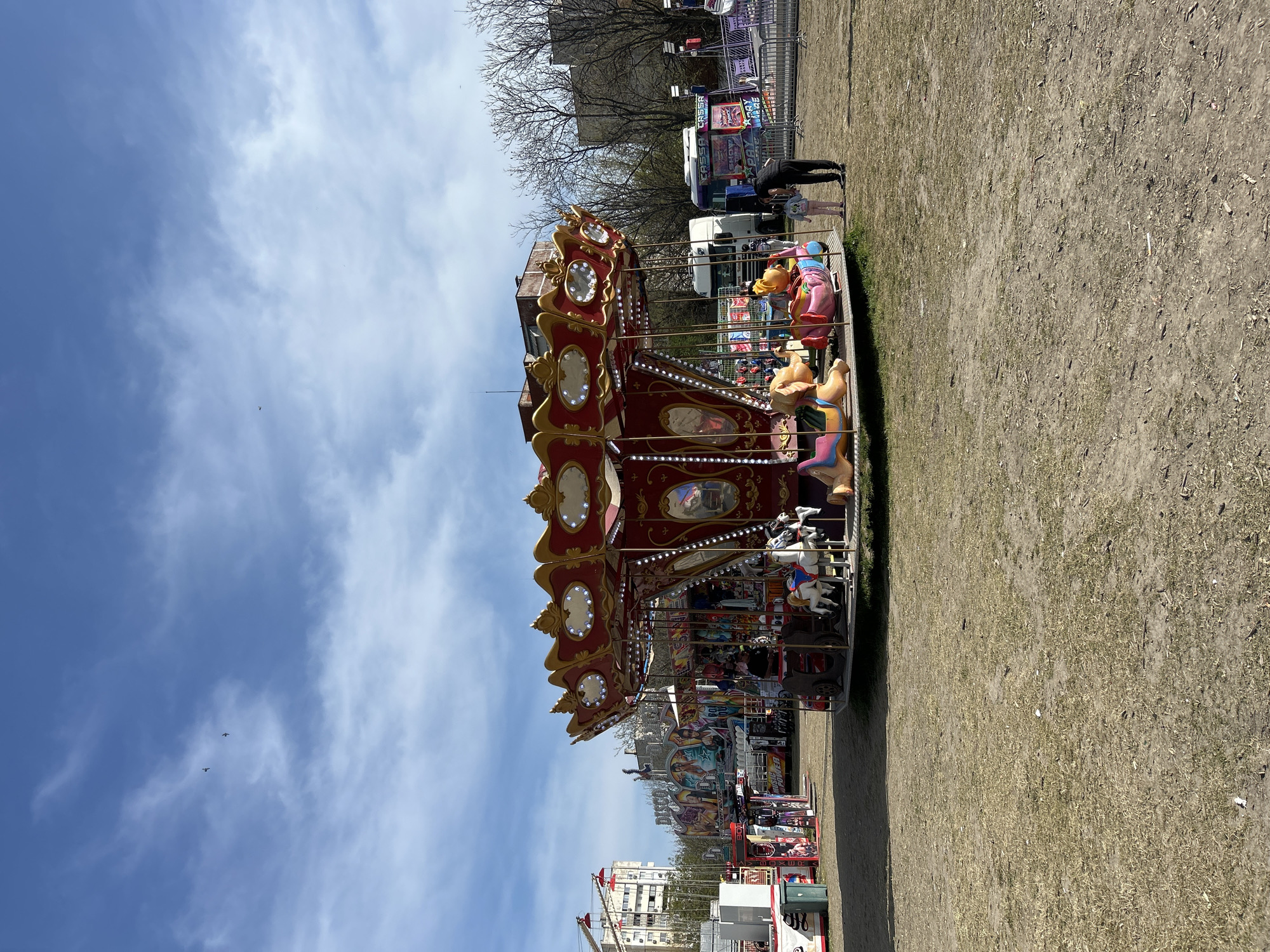 The image captures a vibrant scene at an outdoor fairground, featuring a large, ornate carousel as the central subject. The carousel is predominantly red and gold, adorned with intricate patterns and numerous embedded lights. It features multiple levels of classic animal figures, including various horses and other colorful creatures, ready for riders.
The ground around the carousel is dry and uneven, a mix of dirt and sparse grass, suggesting a temporary setup for the fair. In the foreground, near the carousel's edge, an adult in dark clothing is seen bending towards a small child, who wears lighter attire, possibly assisting them or engaging in conversation.
In the background, behind the carousel, other fairground attractions are visible, including what appears to be a white van or trailer with colorful graphics and some indistinct, brightly colored stalls or rides. A few bare-branched trees stand in the mid-ground, indicating late winter or early spring. Further in the distance, parts of urban buildings can be seen under a bright sky.
The sky is a clear, light blue with scattered wispy white clouds. Two small, dark objects, possibly birds or distant aircraft, are visible high in the sky. The bright lighting suggests it is daytime, likely morning or early afternoon, with pleasant weather conducive to an outdoor event.
The overall scene depicts a lively fair or amusement park atmosphere in Plovdiv, Bulgaria. While most text on distant structures is illegible, some faint letters like "CANDY" or similar are vaguely discernible on the graphics of the white van/trailer.