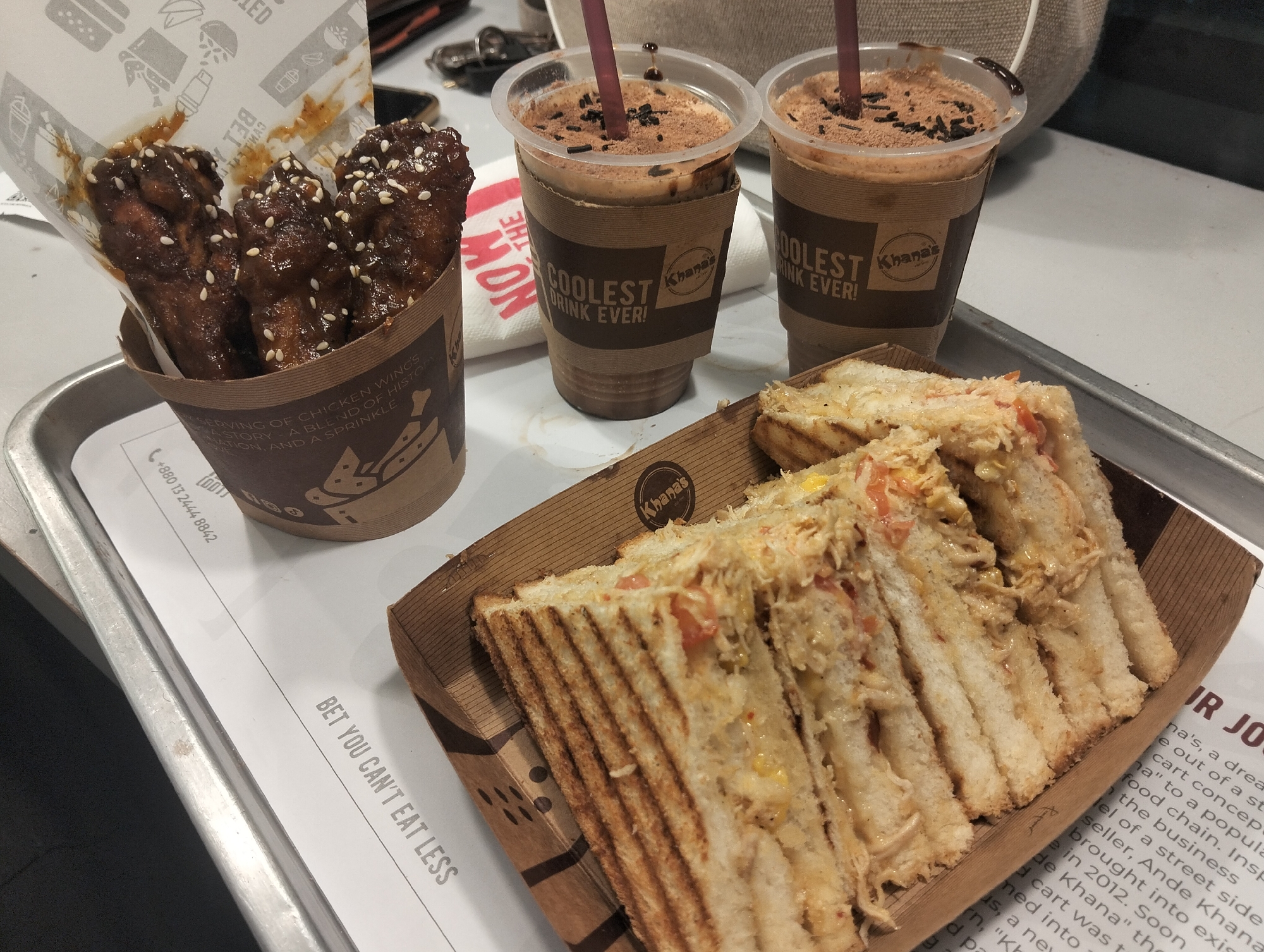 A casual meal from "Khana's," a street-side food chain originating in Dhaka, Bangladesh, is presented on a metal tray, likely within an indoor restaurant or cafe setting.
The meal includes a serving of glazed chicken wings, generously sprinkled with sesame seeds, contained in a brown paper cup. Next to it are two chocolate-flavored iced drinks, topped with cocoa powder and chocolate sprinkles, served in clear plastic cups with branded brown sleeves. The sleeves prominently feature the "Khana's" logo and the slogan "COOLEST DRINK EVER!". A cardboard tray holds several grilled sandwiches, cut into triangular halves, revealing a filling of what appears to be shredded chicken, corn, and diced tomatoes.
The food items rest on a white paper liner detailing the brand's history, mentioning its 2012 origin by Ande Khana as a "street side food chain." The liner also displays a contact number, "+880 13 2444 8842," confirming the Bangladesh context, and the playful phrase "BET YOU CAN'T EAT LESS." A white napkin is visible, partially obscuring the word "THE," along with keys and a blurred bag in the background. The arrangement suggests a ready-to-eat, satisfying meal.