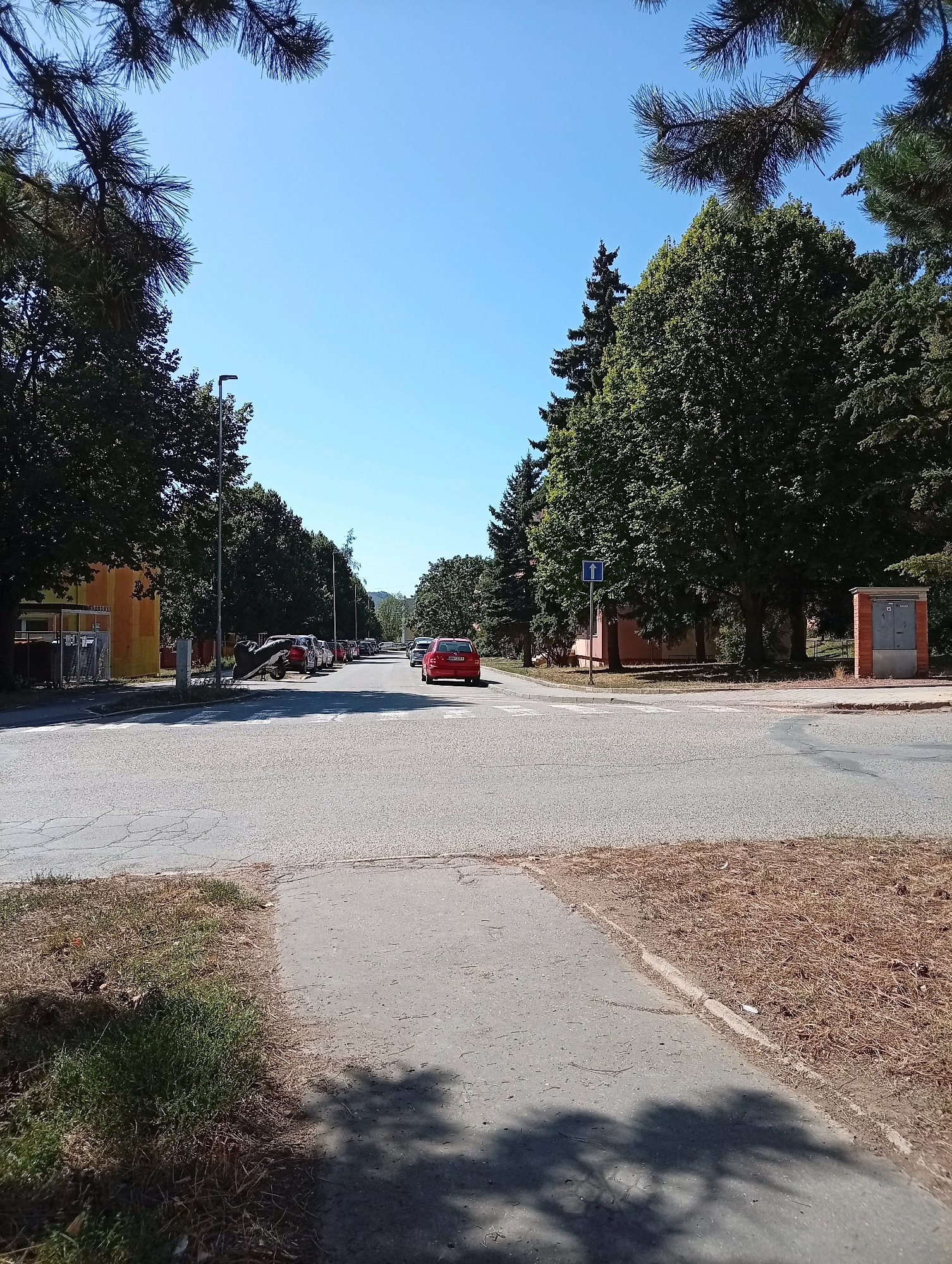 The image depicts a street scene in Tišnov, Czechia, on a clear, sunny day. The viewpoint is from a paved walkway adjacent to a road intersection. In the foreground, the textured asphalt of the walkway is visible, with patches of dry grass and pine needles on the side. The road surface exhibits some cracking.
The intersection itself features a pedestrian crossing. Several parked cars are lined up along the right side of the road, and a red car is in motion on the intersecting road. A street lamp stands on the left side of the road, and a blue traffic sign with a white arrow pointing upwards is mounted on a pole near the intersection.
The environment is characterized by mature trees with dense green foliage lining both sides of the street. On the left, a building with an orange and yellow facade is partially visible. On the right, there's a small brick structure that appears to be an electrical utility box. The sky is a clear, bright blue. The scene suggests a residential or suburban area.
