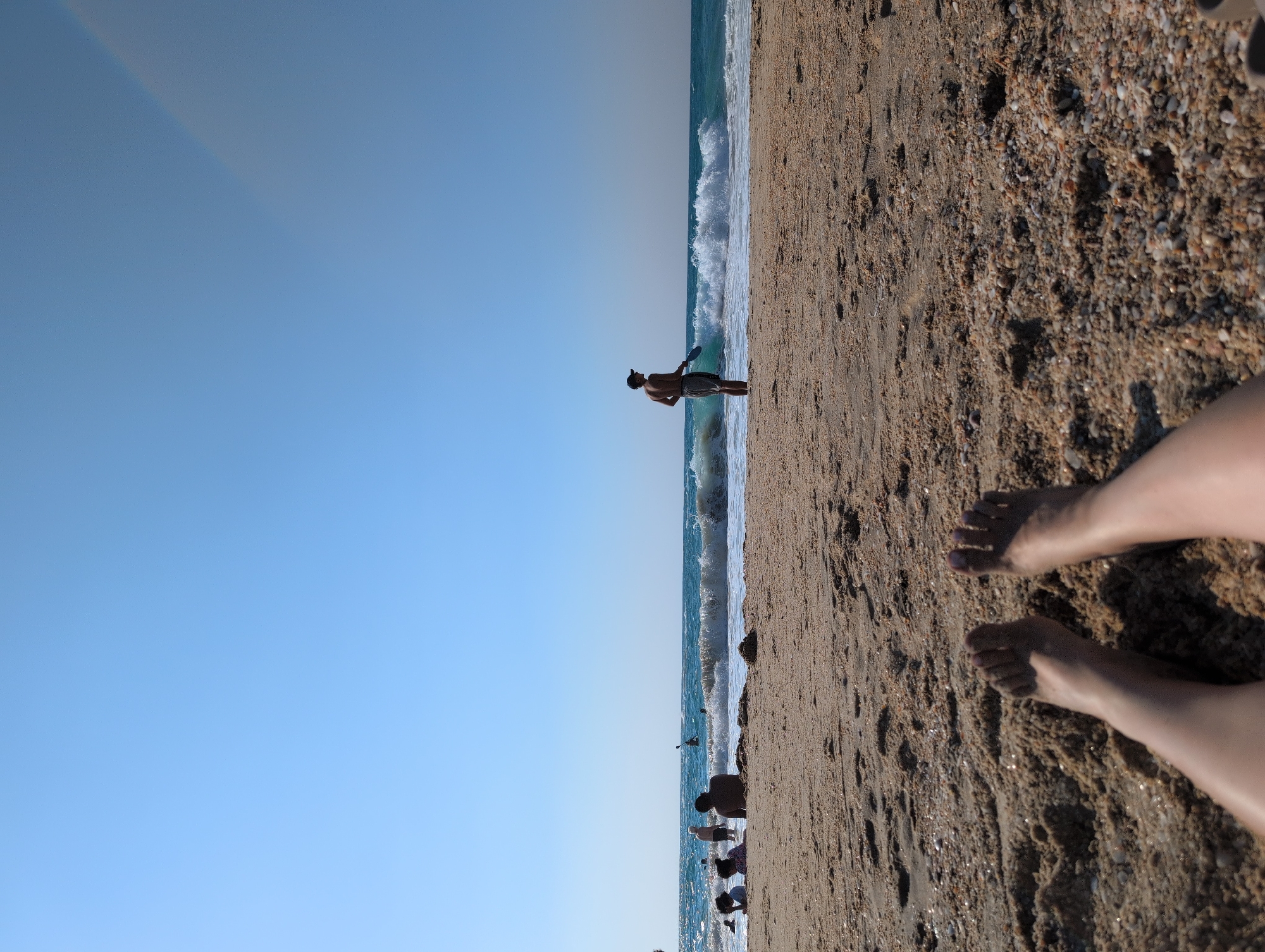 The image shows a beach scene at Palmachim, Israel. The foreground features a close-up of a person's bare legs and feet lying on the sand. The sand is coarse with small pebbles and shells. In the midground, several people are visible in and near the water. Some are wading, while others appear to be standing or sitting on the beach. Waves are breaking in the ocean, creating white foam. In the background, a person stands by the water's edge holding a paddle-like object. The sky is clear and blue with a slight haze near the horizon. The overall impression is a sunny day at the beach.