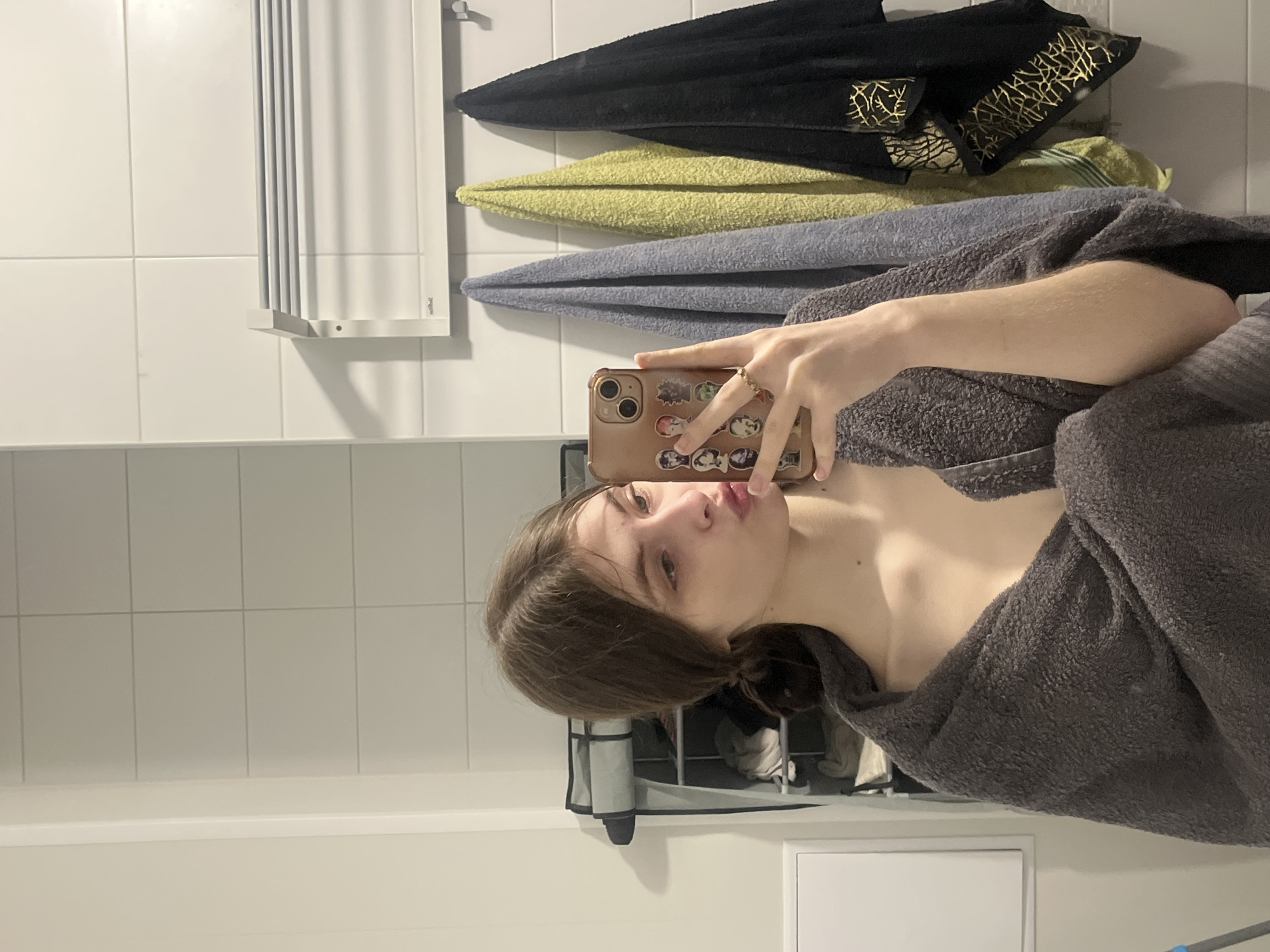 A person is seen taking a mirror selfie in what appears to be a bathroom. The subject has brown hair and is wearing a dark grey, fluffy robe or towel. Their face is partially visible, with lips puckered in a pout. They are holding a beige/light brown smartphone, adorned with numerous small, colorful cartoon or anime character stickers on its case. A ring is visible on one of the person's fingers.
The background consists of white tiled walls, typical of a bathroom. To the left, a white, accordion-style wall-mounted drying rack or towel holder is visible. To the right, above the person's shoulder, several neatly folded towels are hanging, including a black one with a distinctive gold pattern, a light green one, and a light blue one. Below the tiled wall, a white surface, possibly a shelf or counter, runs across the frame. In the lower part of the image, behind the person, a grey and black collapsible laundry hamper or storage unit can be seen. A white access panel or vent is also visible on the lower wall. The lighting suggests an indoor setting, likely a home or hotel in Saint Petersburg, Russia, as per the provided context. No discernible text is visible in the image.