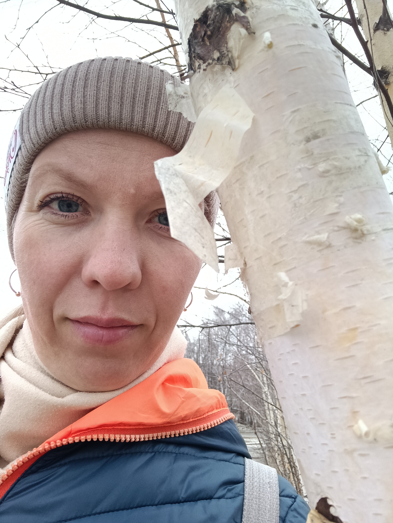 A selfie shows a woman standing next to a birch tree. She wears a beige beanie, a light beige scarf, and a dark blue puffer jacket with an orange hood liner. The bark of the birch tree is peeling. The background is blurred, showing bare trees, suggesting it might be autumn or winter. She likely took this picture in a park or wooded area. While the city is not explicitly shown in the image, it's stated this image was taken in the city of Chaikovsky, Russia.