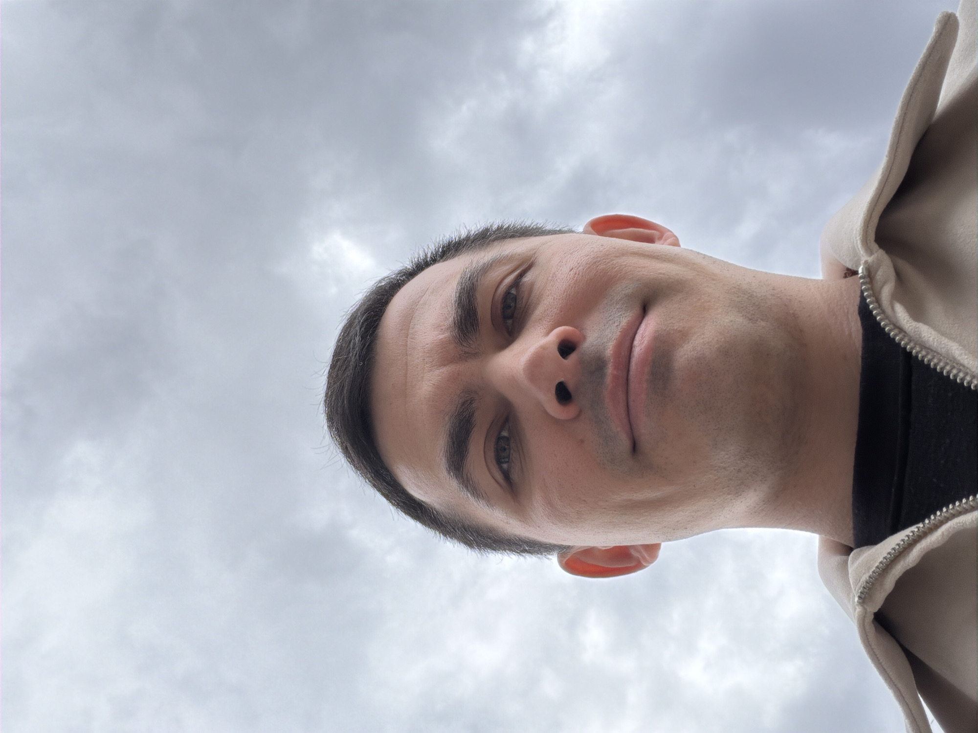 A man is looking down at the camera from a low angle. He has dark hair and light skin. He is wearing a zipped jacket with a black collar and a white/cream-colored outer layer. The background is a cloudy sky. It's impossible to determine the exact location of Izhevsk, Russia from this image.
