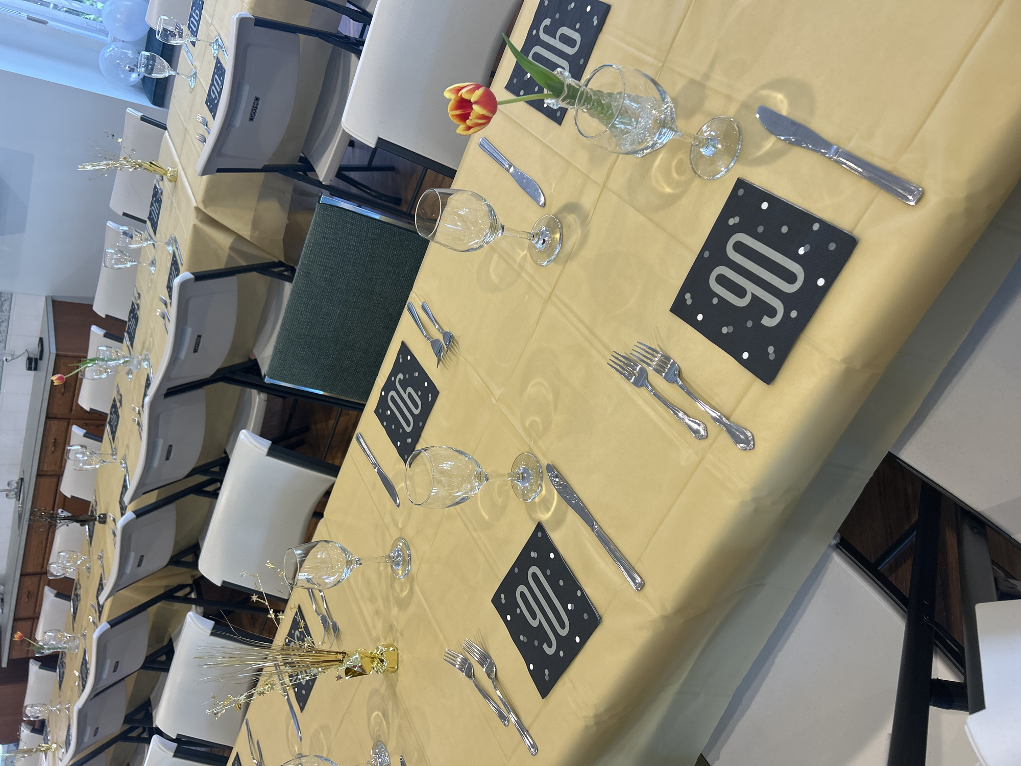 The image depicts a dining table set up for a 90th birthday celebration. The location is difficult to determine from the image; there is no clear indication this is Kelowna, Canada. A long table is covered with a light gold tablecloth. Each place setting includes a wine glass, a knife, two forks, and a black square card with "90" printed in silver. Some place settings also have a small vase with a single tulip. Other centerpieces include small gold-colored decorations. White folding chairs are arranged around the table. The background shows part of a room with white walls and some cabinetry. The lighting is bright.