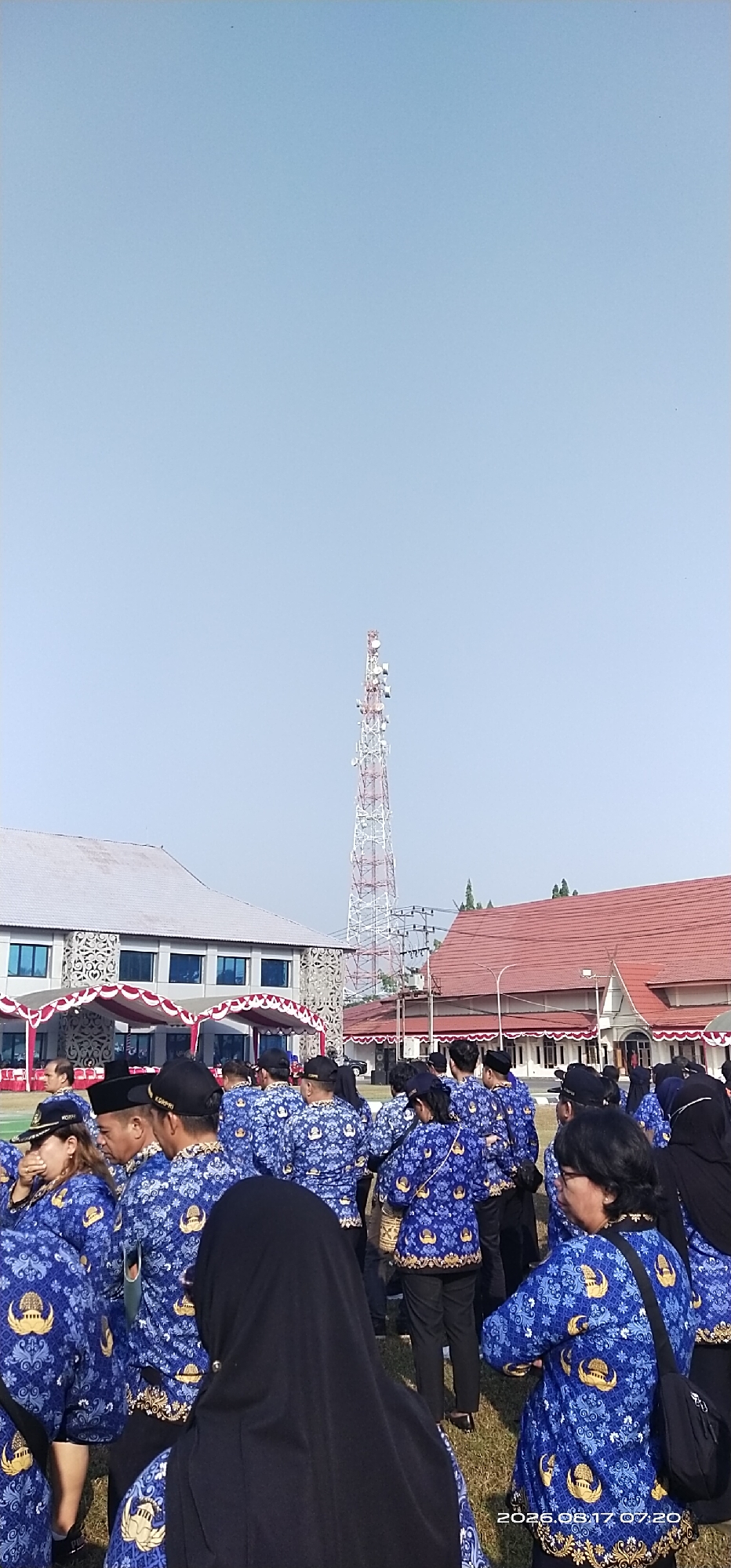 A group of people in matching blue shirts with a gold emblem are gathered outdoors. They are wearing black hats and some are wearing headscarves. In the background, there are buildings and a tall cell tower. The ground is covered in grass. The scene appears to be a formal gathering or event. The date stamp on the image indicates August 17, 2026, at 07:20.